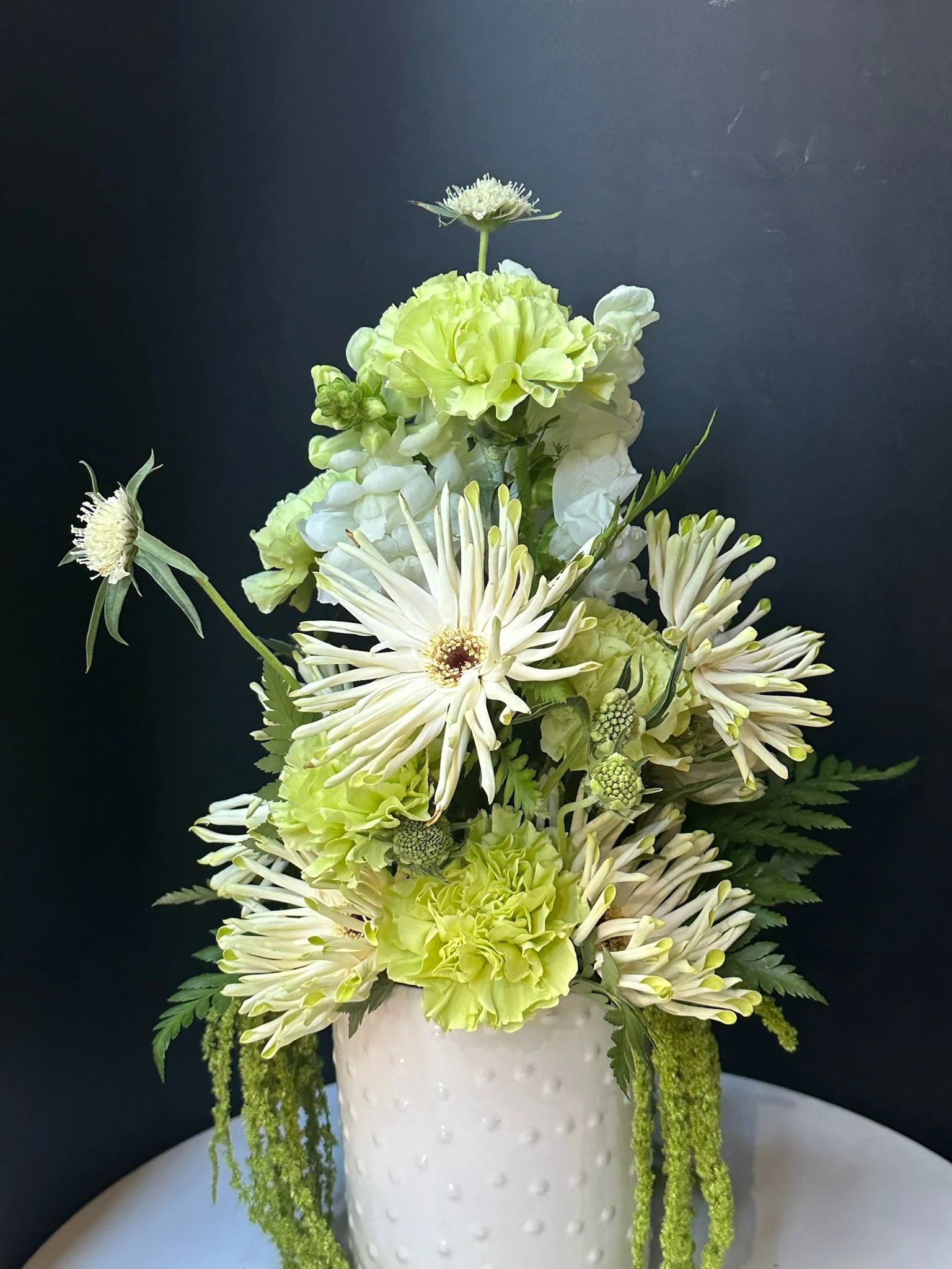 A bouquet of white and green flowers in a white textured vase against a dark background.