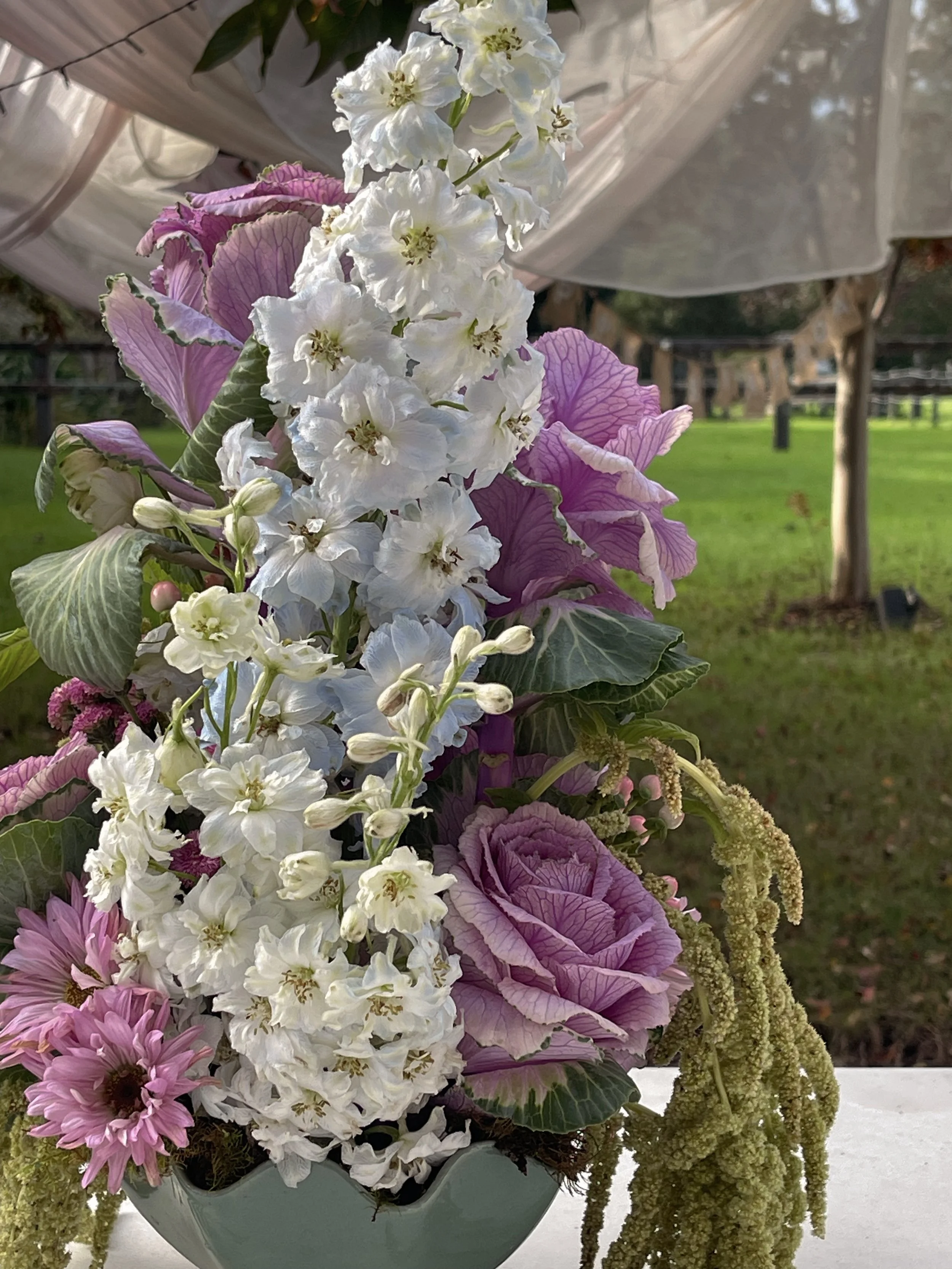 A floral arrangement in a green vase featuring white, pink, and purple flowers with hanging amaranthus, set outdoors with a blurred background of grass and trees.
