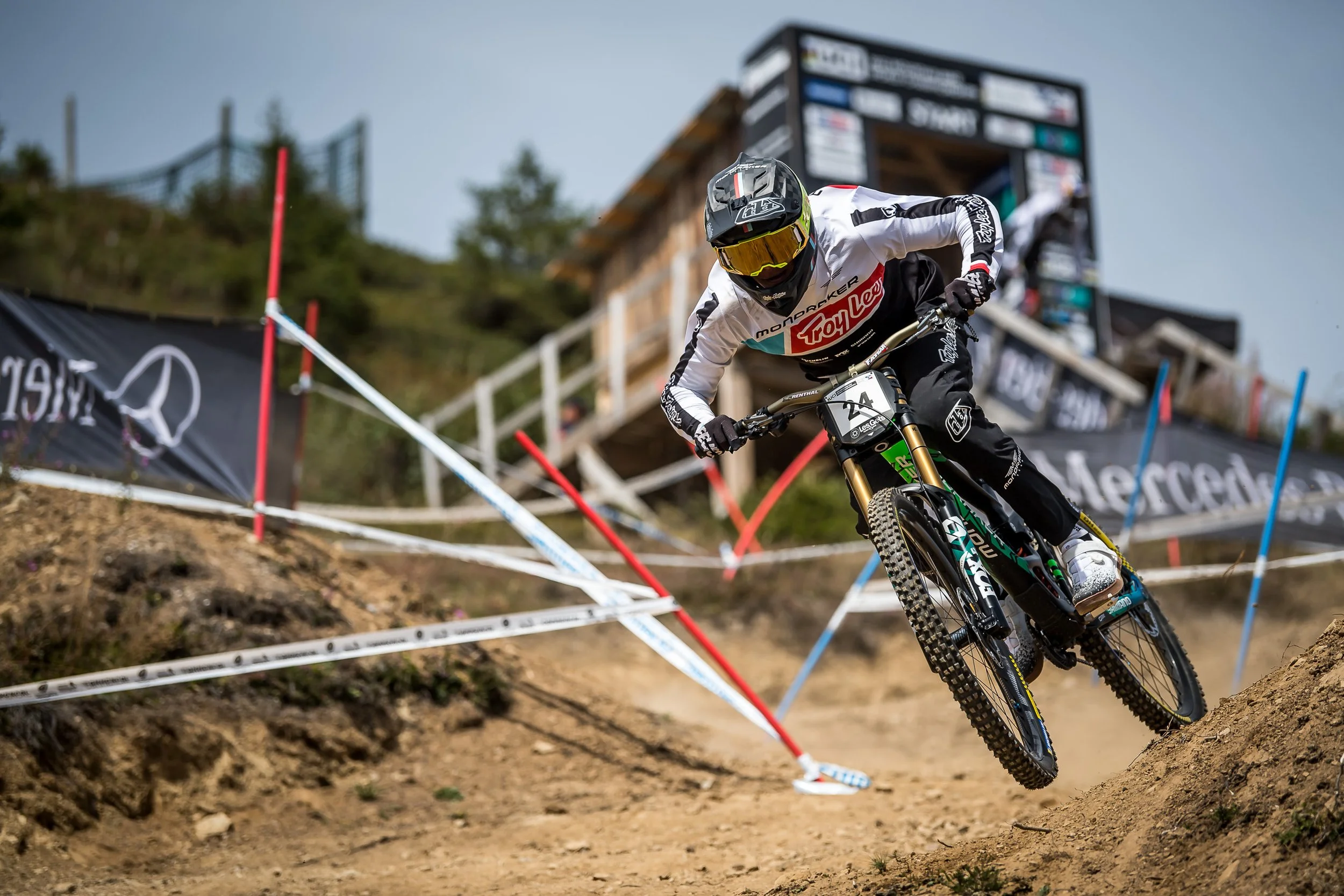 A mountain biker wearing a white jersey with red and black accents, black pants, a helmet with goggles, and gloves, speeds down a dirt trail during a race. The rider is leaning into a turn with a focused posture. In the background, there are sponsor banners, a wooden structure, trees, and a large digital scoreboard.