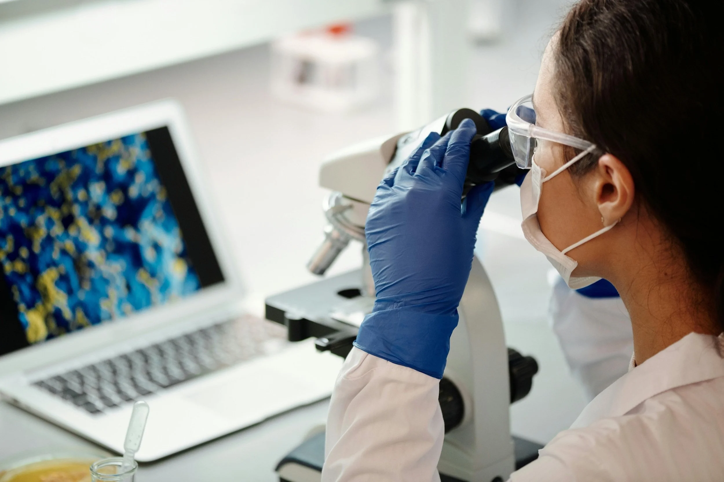 A scientist looking through a microscope in a laboratory, wearing safety goggles and a face mask, with a computer monitor displaying colorful data in the background.