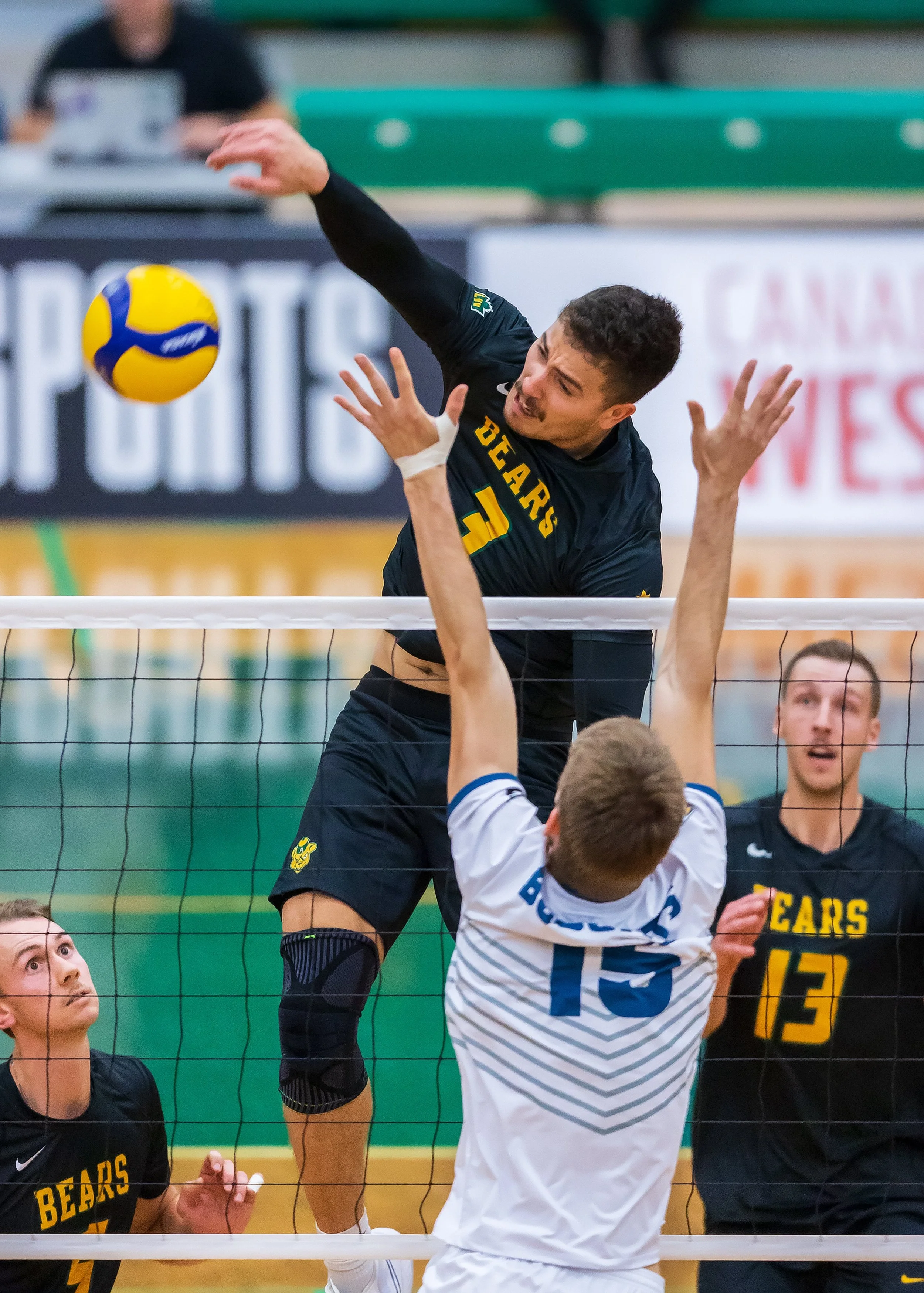 A volleyball player in a black jersey with yellow and green lettering spikes the ball over the net while players in white jerseys attempt to block. The scene is indoors with green bleachers in the background.