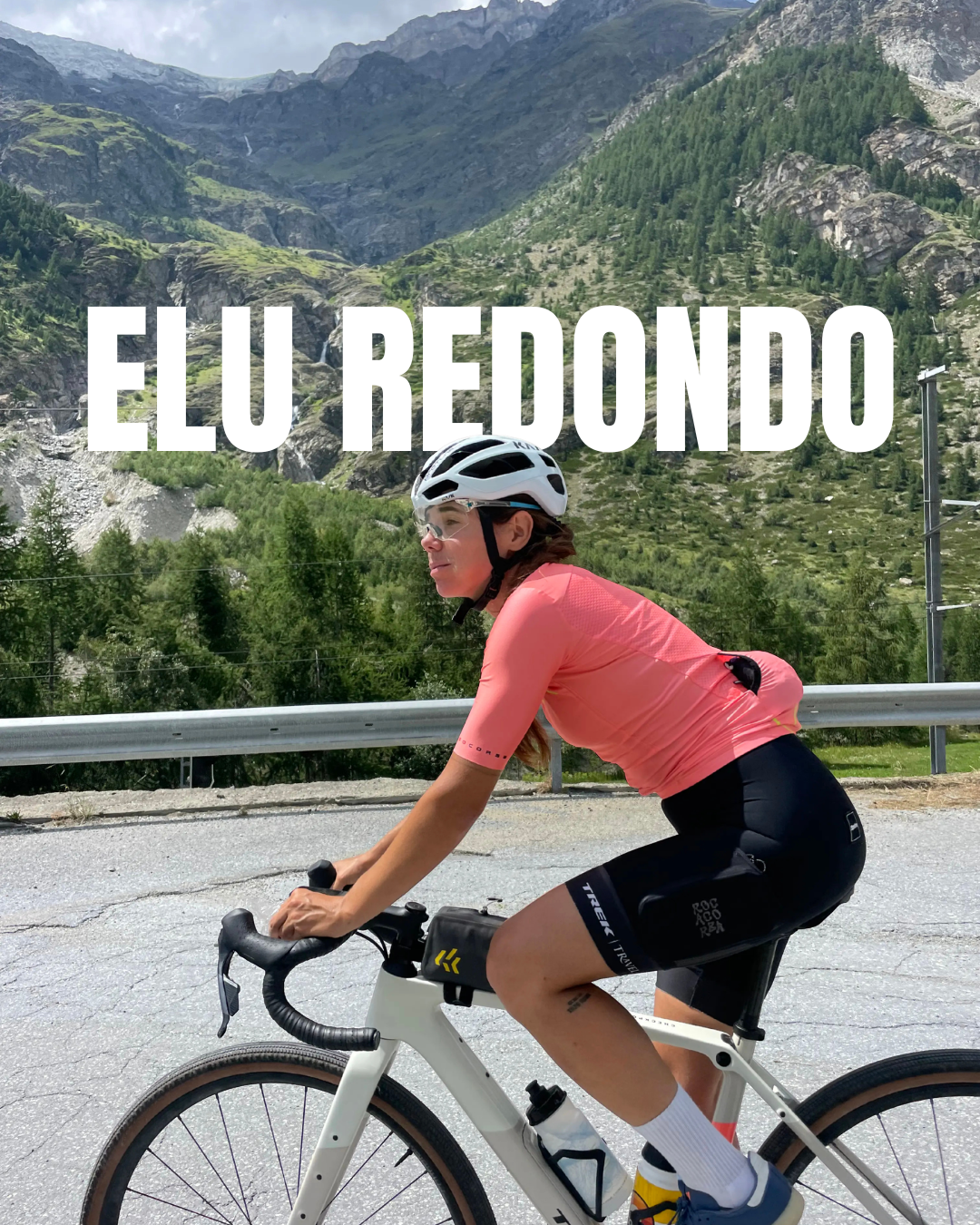 A woman riding a road bicycle along a mountain road with lush green hills and mountains in the background, wearing a pink cycling jersey, black shorts, white helmet, sunglasses, and white socks.