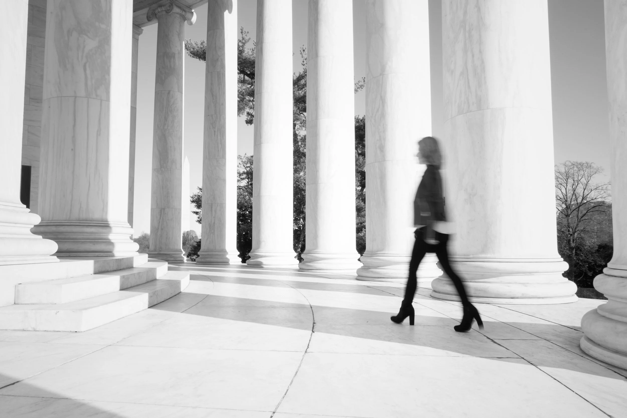 Black and white photo of a person walking past large marble columns on steps outside a government building.