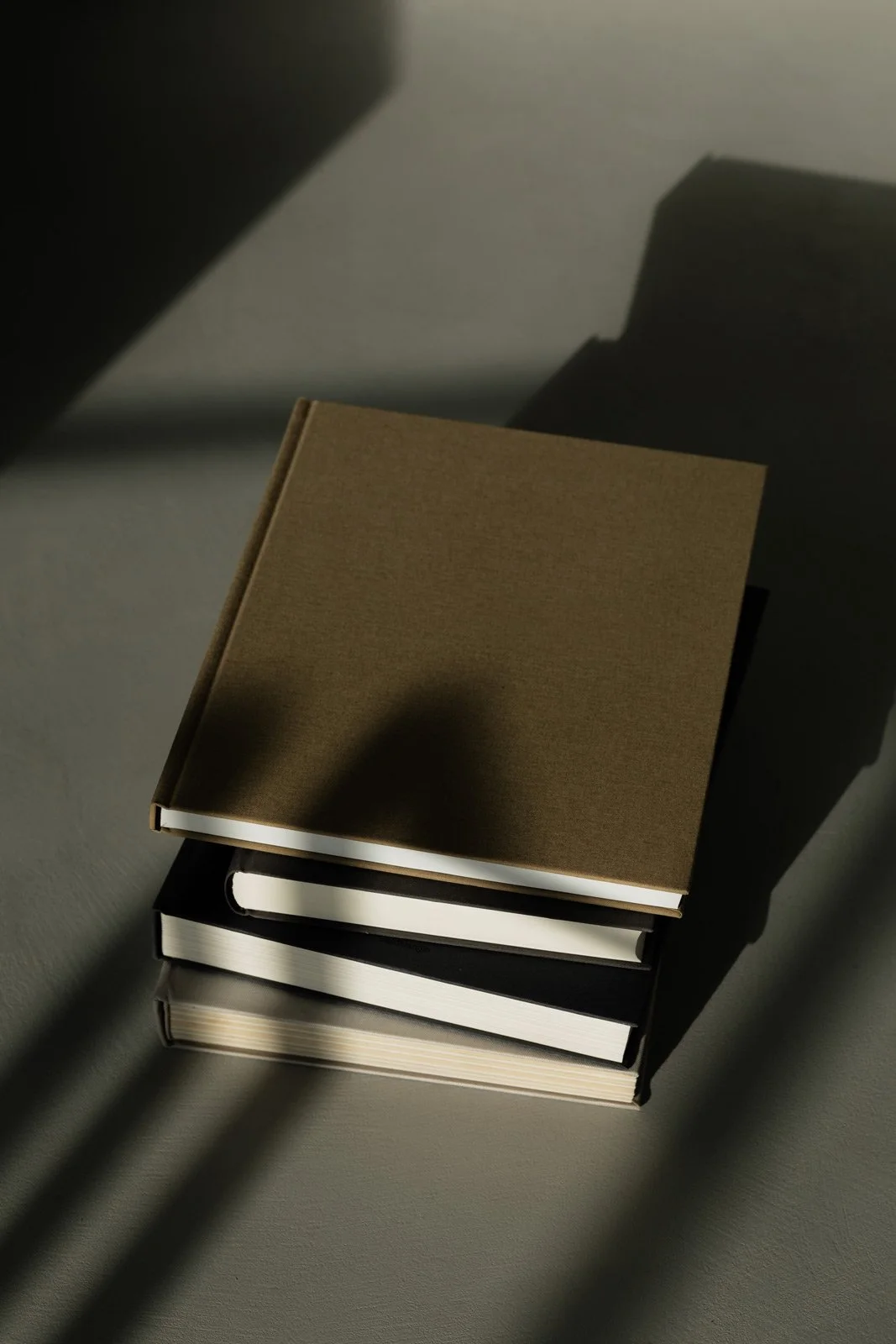 Stack of three books with a plain brown cover on top, placed on a gray surface with shadows cast by window blinds.
