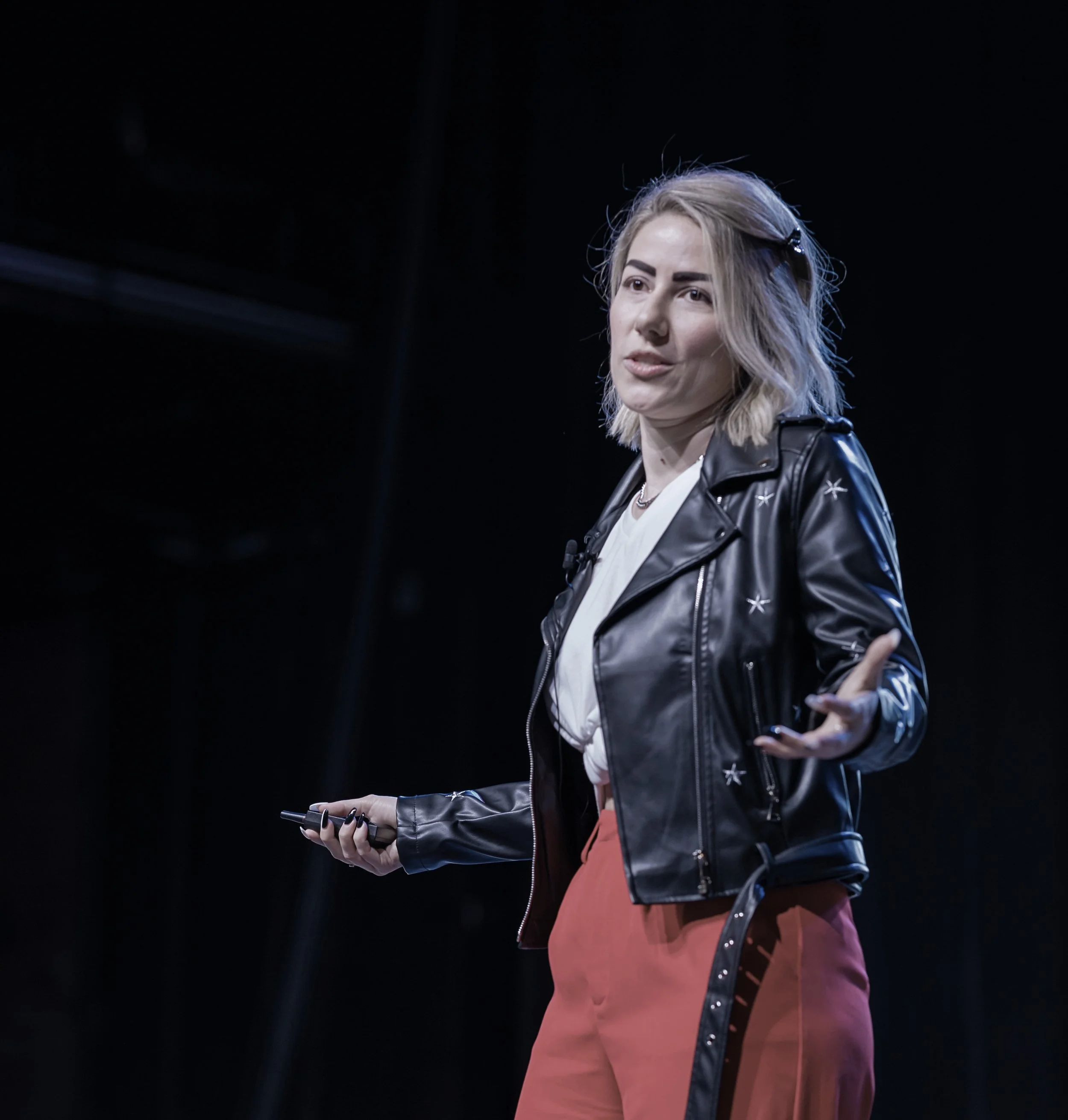 Young woman with blonde hair, wearing a black leather jacket, standing against a dark background, holding a phone in her hand, engaged in conversation.
