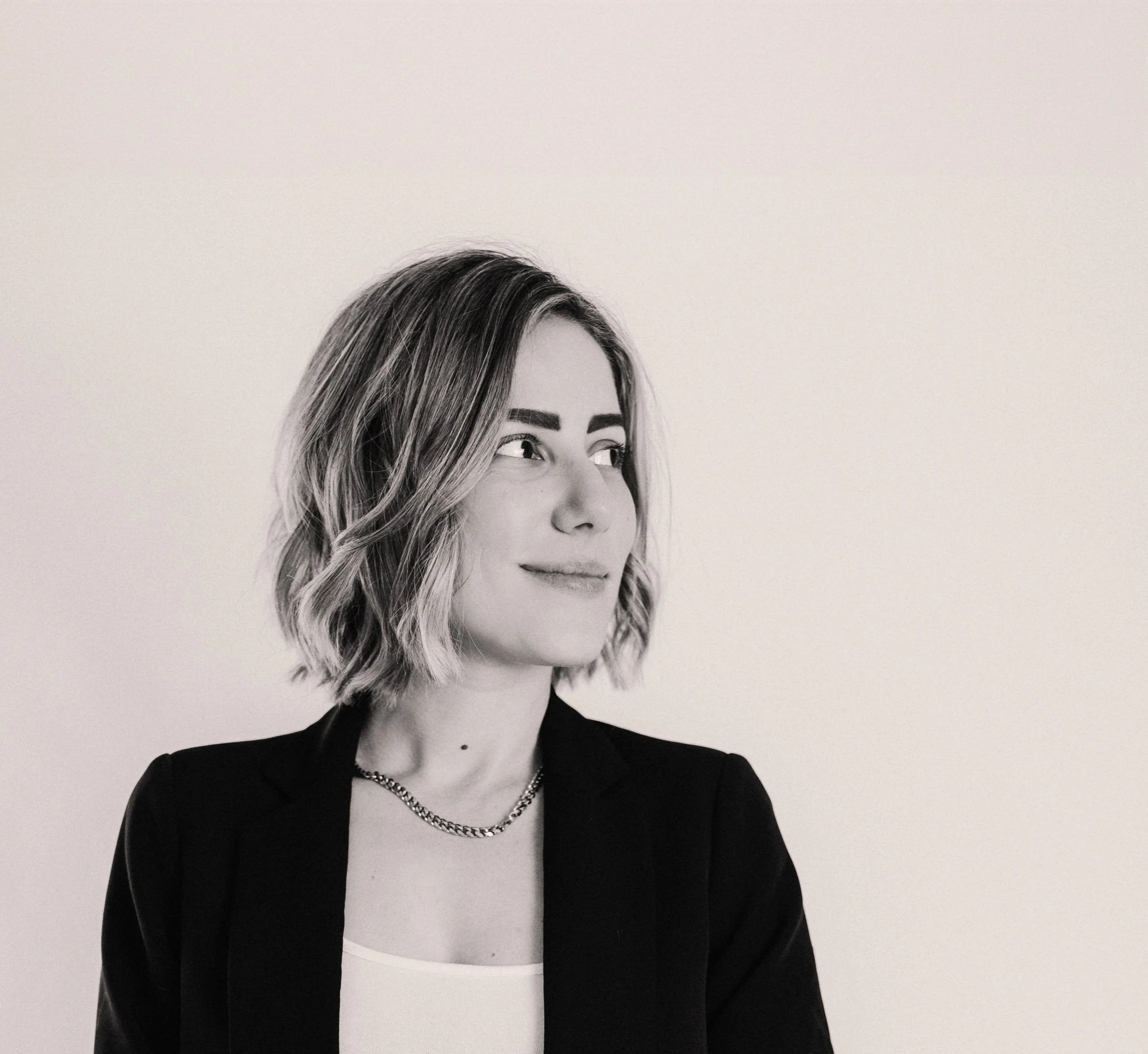 Black-and-white portrait of a woman with medium-length hair, wearing a black blazer, a white top, a chain necklace, and subtle makeup, standing against a plain light-colored background.