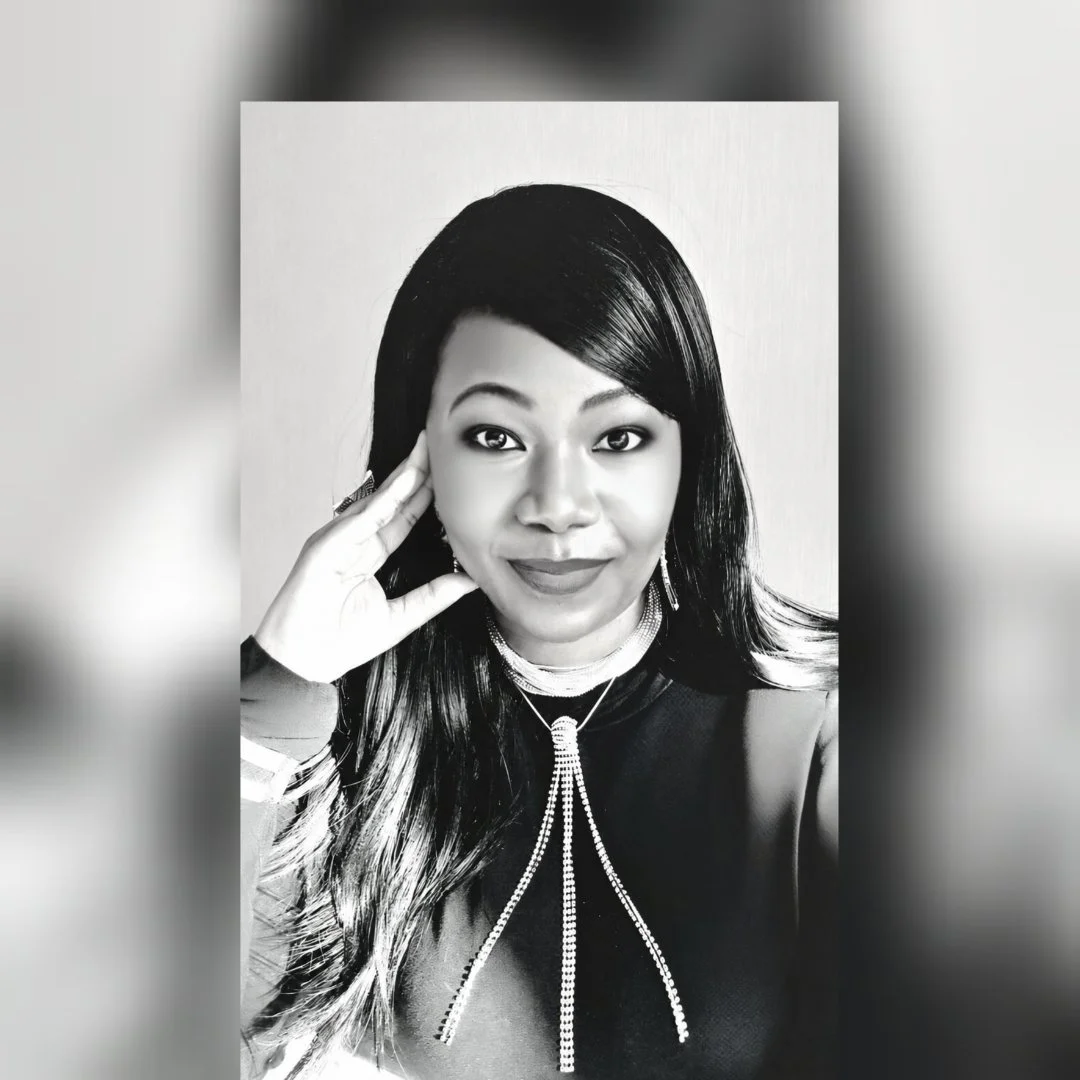 Black and white photo clinical psychologist Maropeng Ralenala with long dark hair, wearing a necklace with a pendant, and a top with a decorative ribbon. She is smiling slightly and resting her hand near her face.
