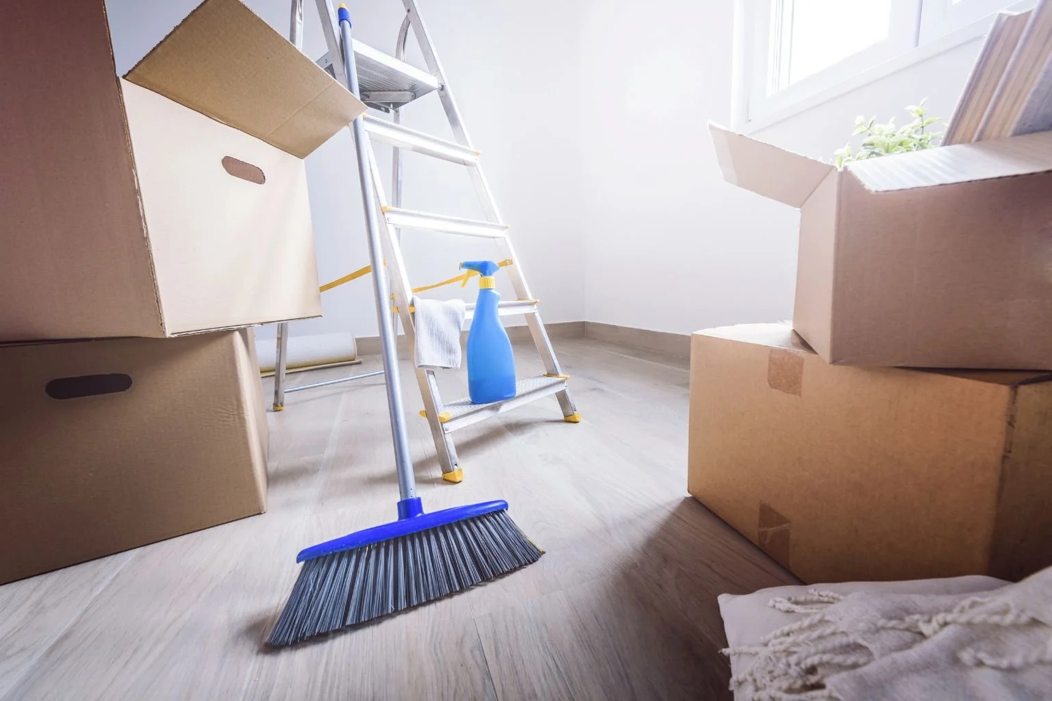 A room filled with moving boxes, a ladder, a broom, and cleaning supplies, indicating a cleaning or moving activity.