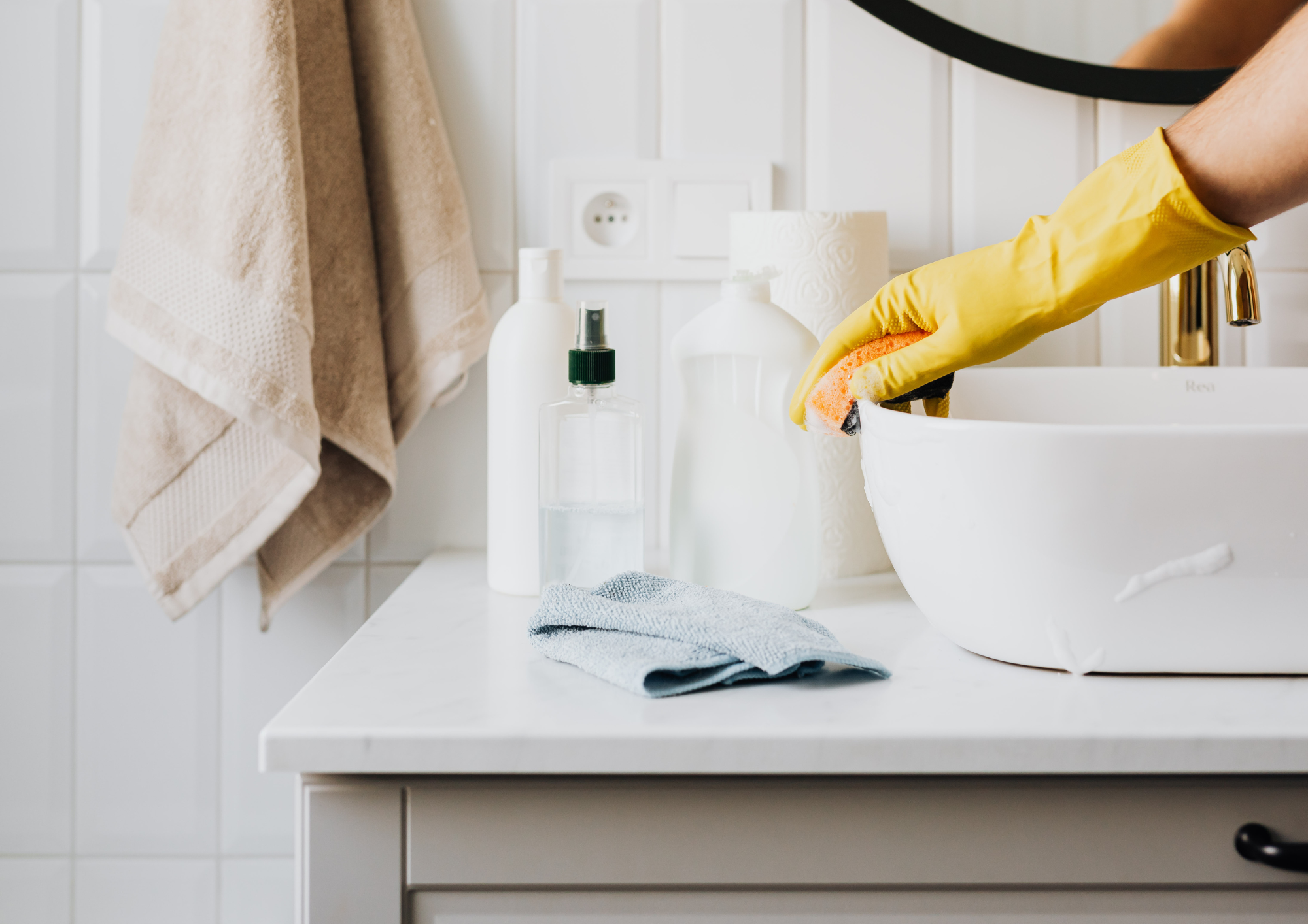 Close-up of a bathroom countertop with a person wearing yellow cleaning gloves scrubbing a white sink with an orange sponge. The countertop has bottles of cleaning supplies and a roll of paper towels. Towel hanging on the wall and an electrical outlet are also visible.