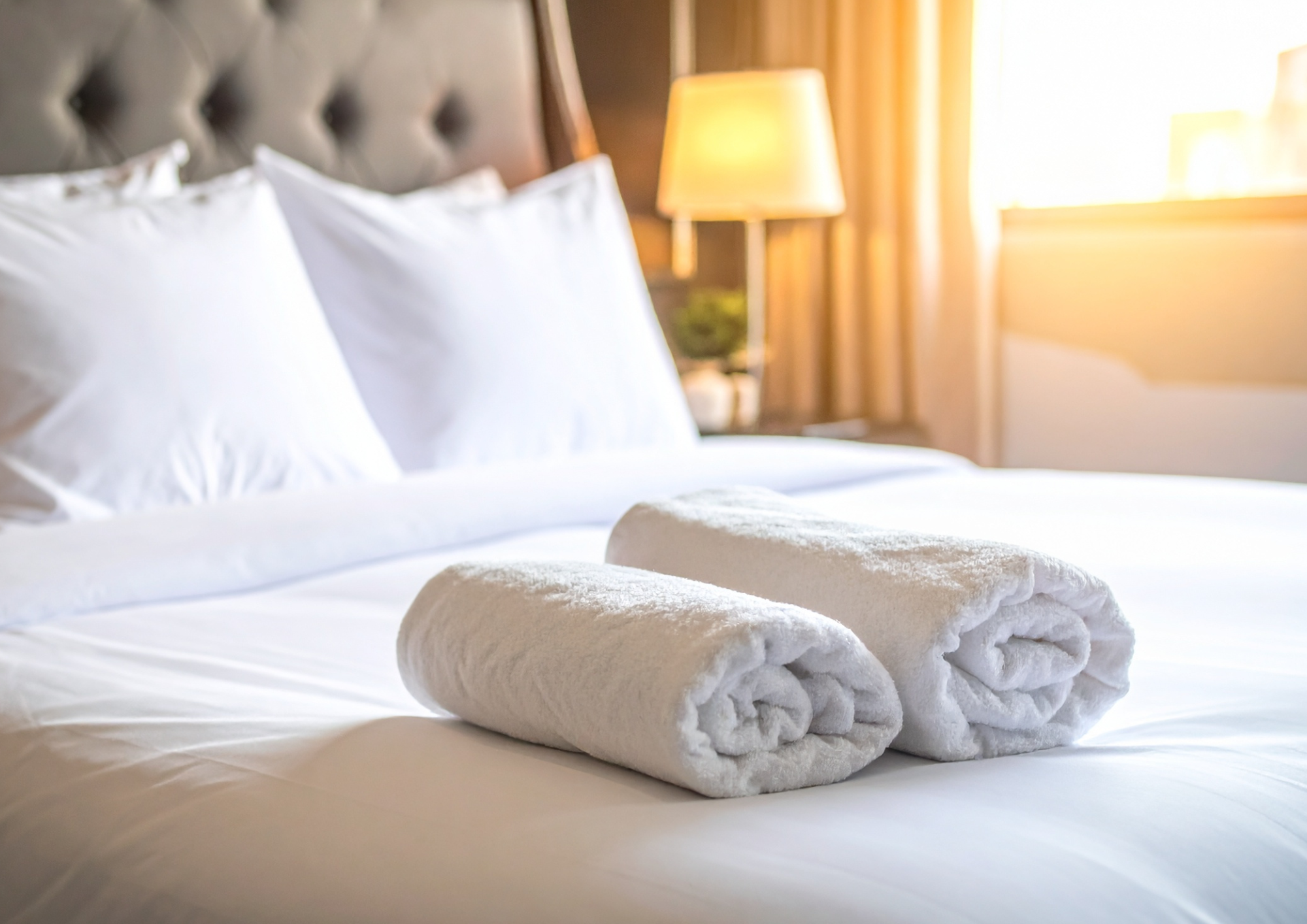 Folded white towels on a bed in a hotel room with white pillows and a bedside lamp.