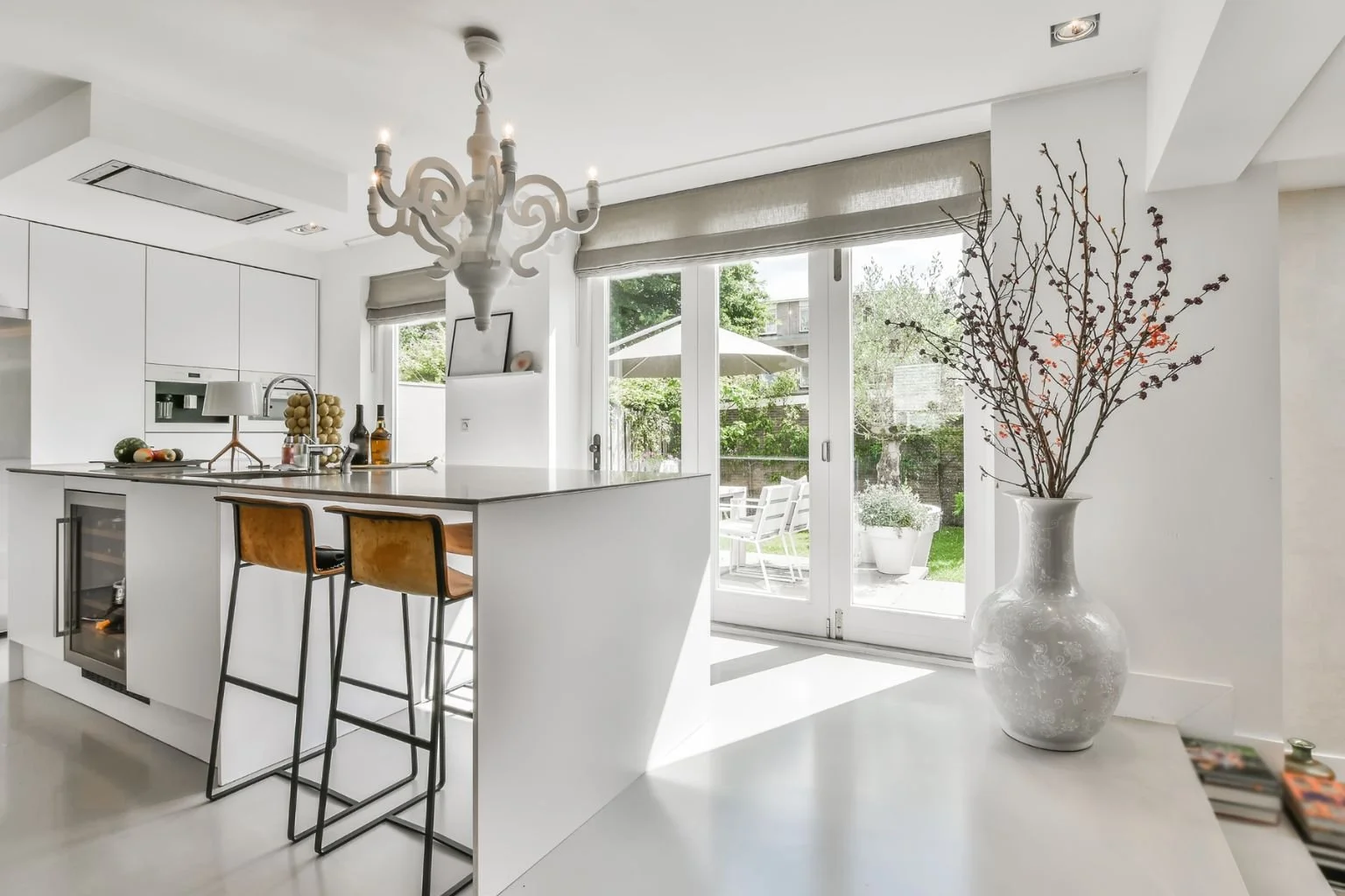 Modern kitchen with white cabinets, a white kitchen island, two tan bar stools, a large white vase with branches, and large sliding glass doors leading to a patio with outdoor furniture.