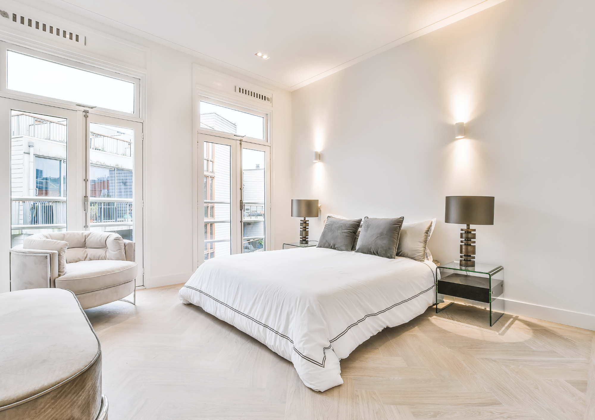 Modern bedroom with large windows, white walls, a white bed with black trim, two gray pillows, two black lamps on glass side tables, and a white armchair with a rounded back in the corner.