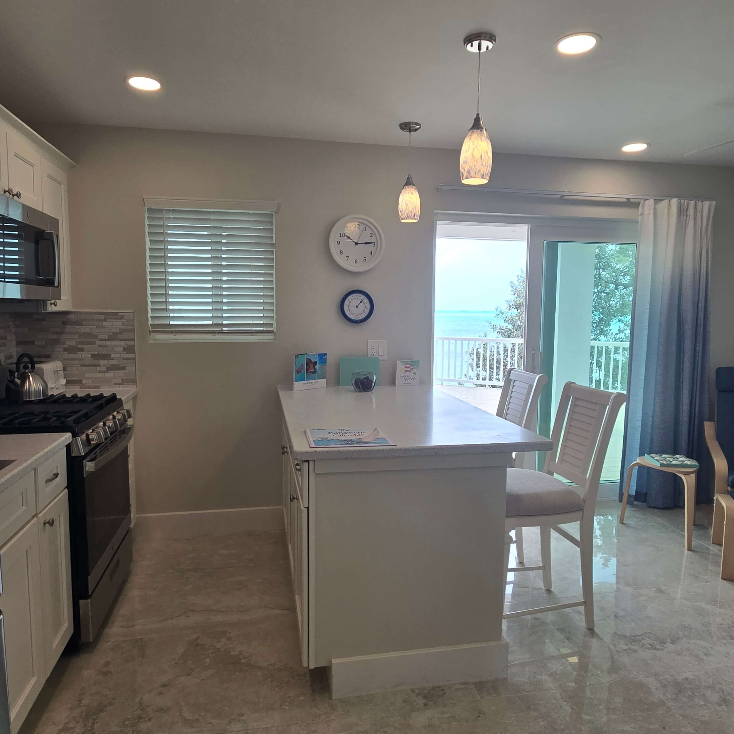 A kitchen and dining area with a white kitchen island, white chairs around a white table, a wall clock, a window with blinds, and pendant lighting, with an open door revealing a deck and outdoor scenery.