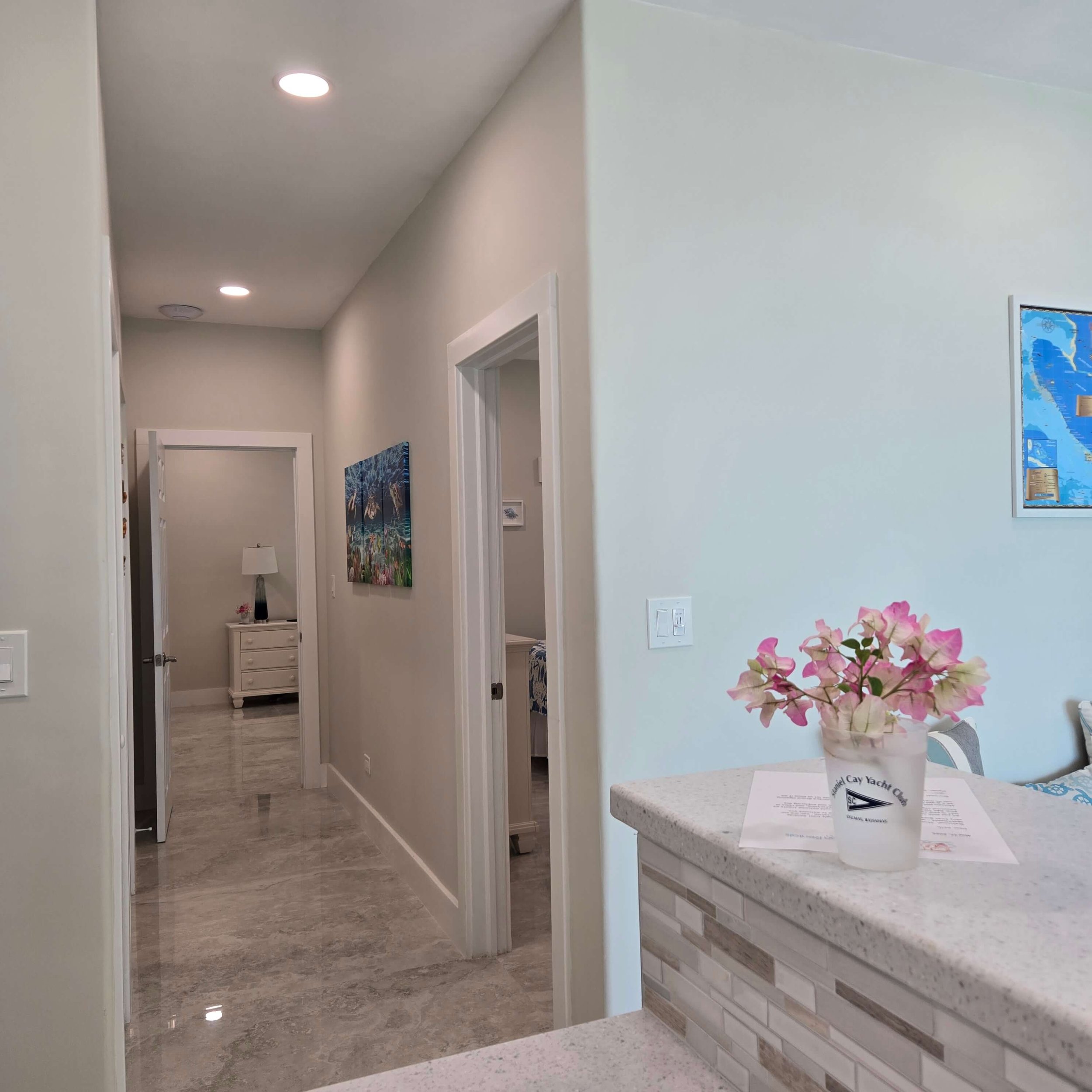 A hallway with beige walls, marble tile flooring, and ceiling lights. Artwork decorates the walls, and there is a small table with a lamp near the end of the hall. On the right side, part of a light-colored kitchen counter with a vase of pink flowers