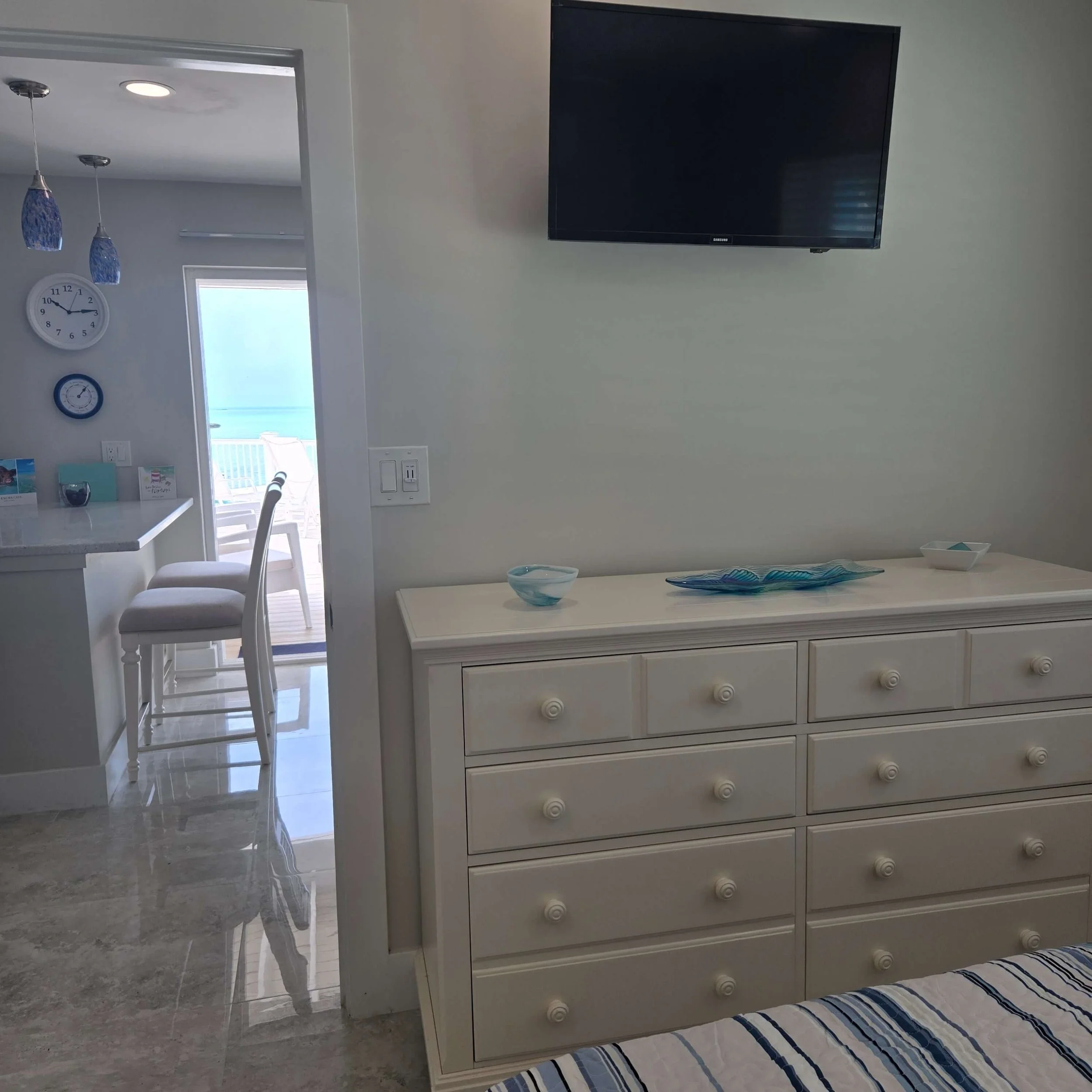 Living room with a wall-mounted television, a white dresser with drawers, a glass bowl, and a striped bed in the foreground. A doorway leading to a balcony with view of the ocean outside, and a clock on the wall inside.