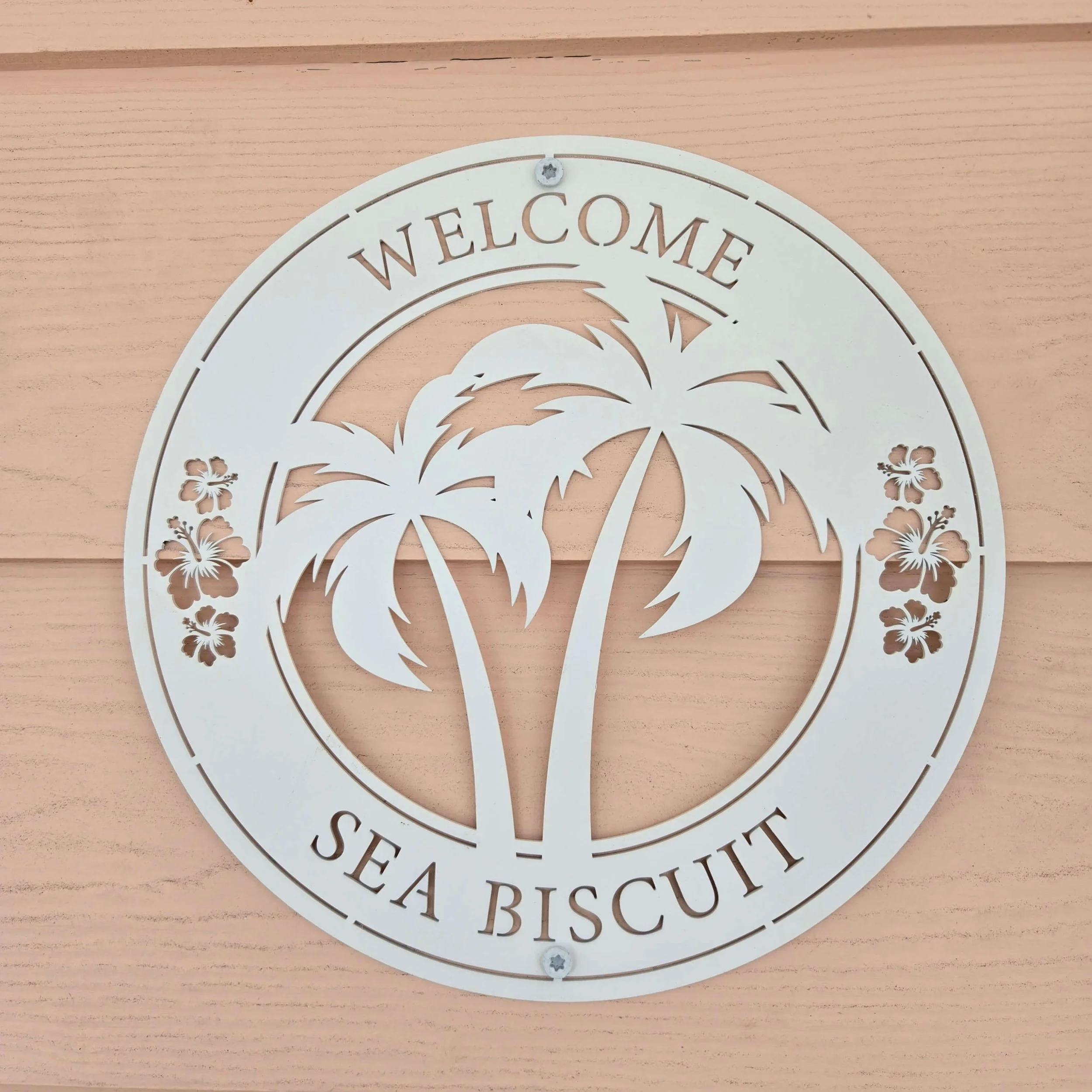 A white decorative sign with the word "WELCOME" on top, featuring a stylized palm tree and hibiscus flowers, mounted on a beige wooden background.