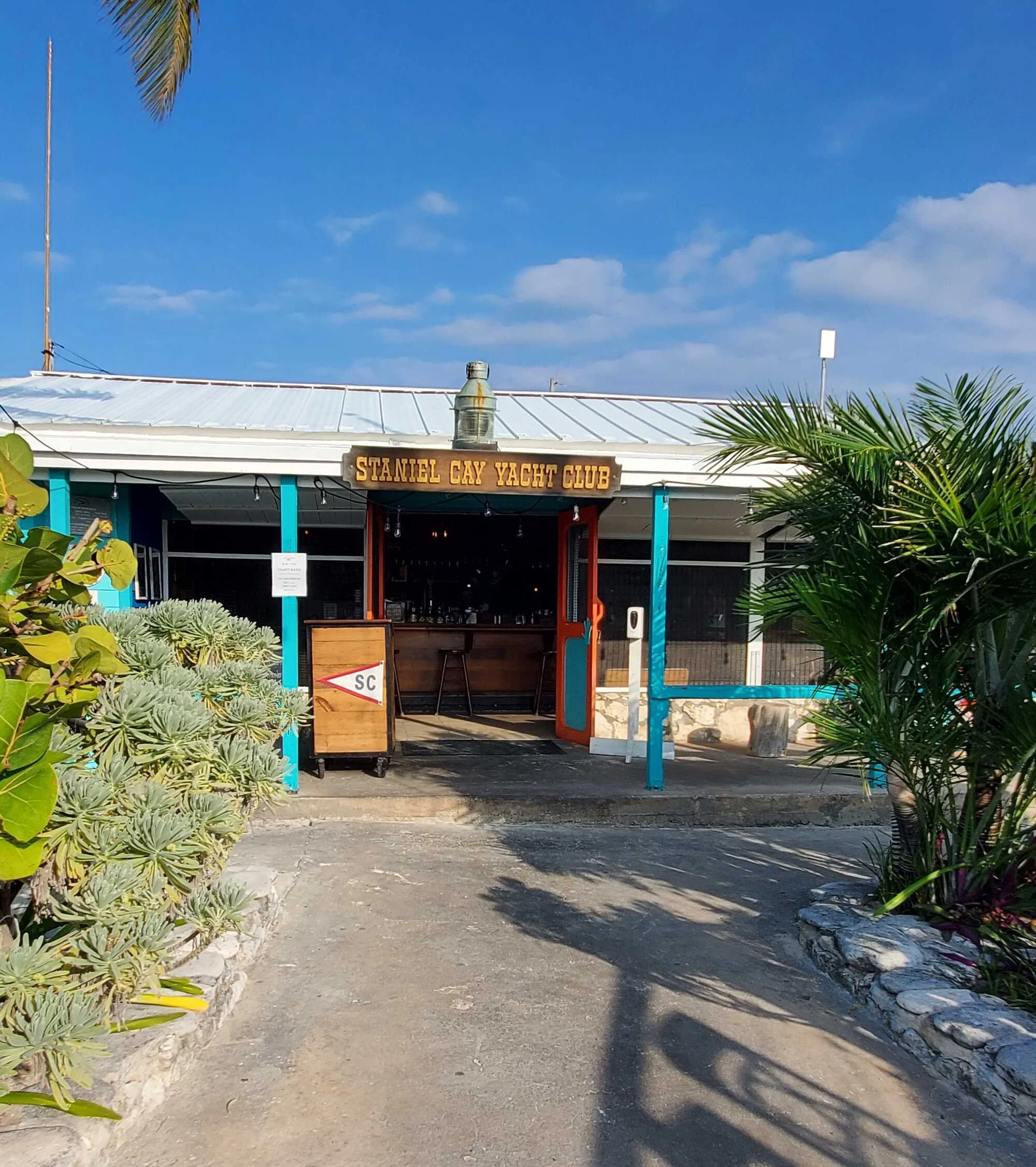 Sign for Stanley Cay Yacht Club in front of a building with a white metal roof, surrounded by tropical plants and bright blue sky with some clouds.