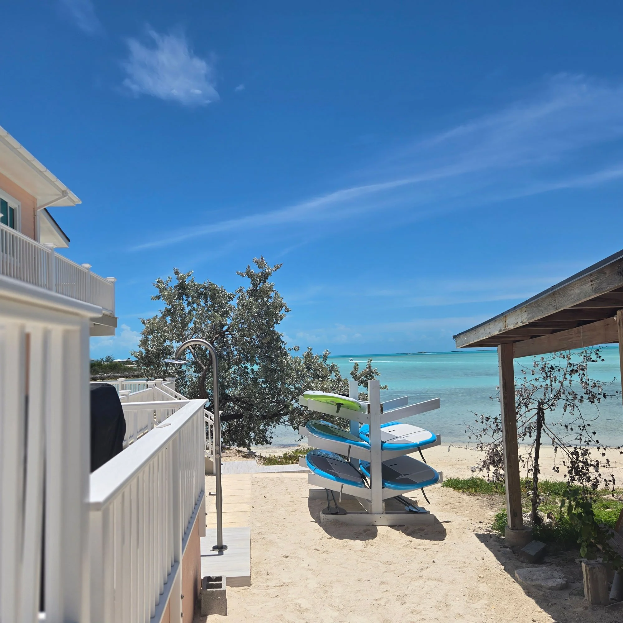 View of Seaside with a white fence, blue skies, and ocean in the background, with SUPs on a rack and a beach shower for guests to use.