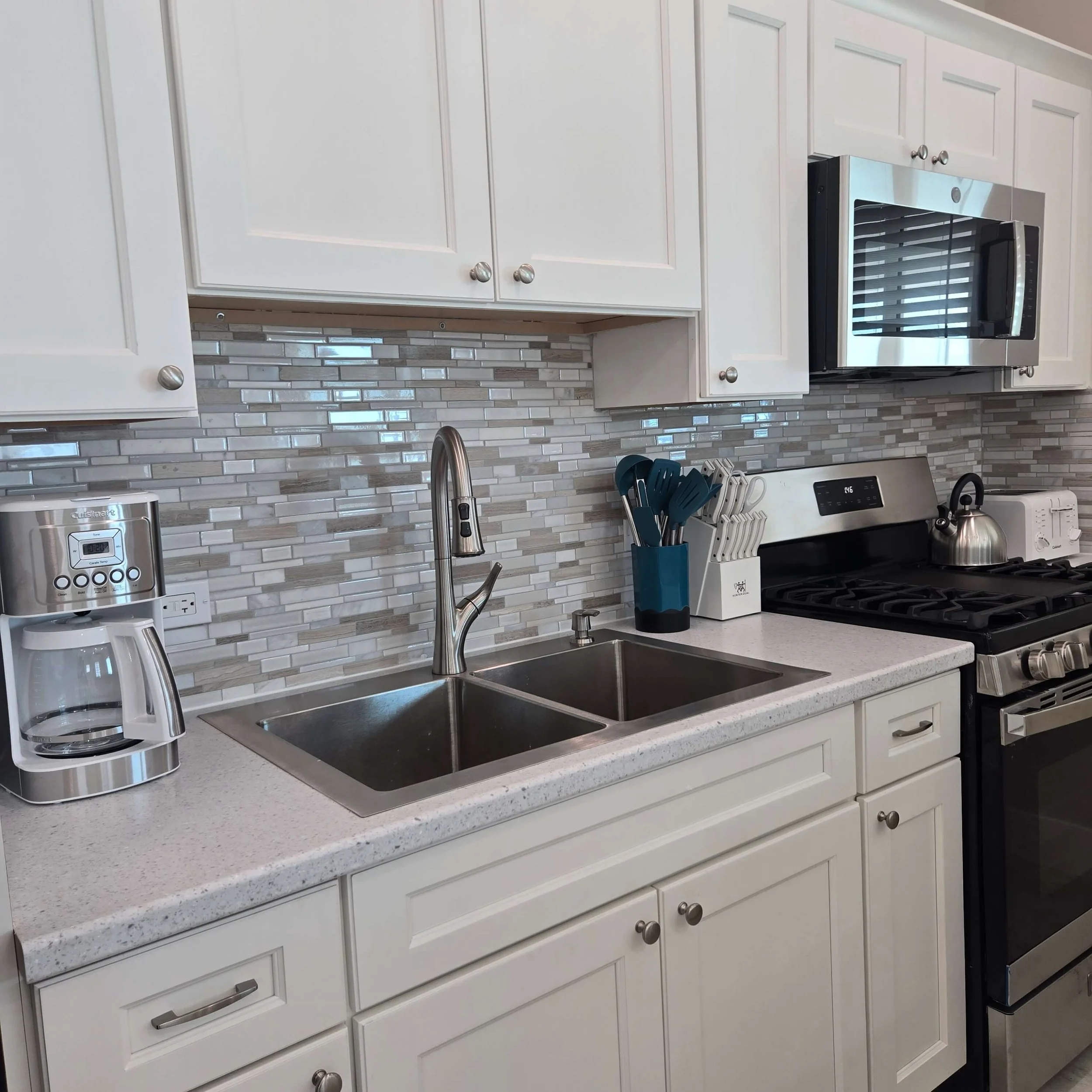 Modern kitchen with white cabinets, gray speckled countertop, stainless steel appliances, and a marble tile backsplash, including a coffee maker, utensils, and a stove.