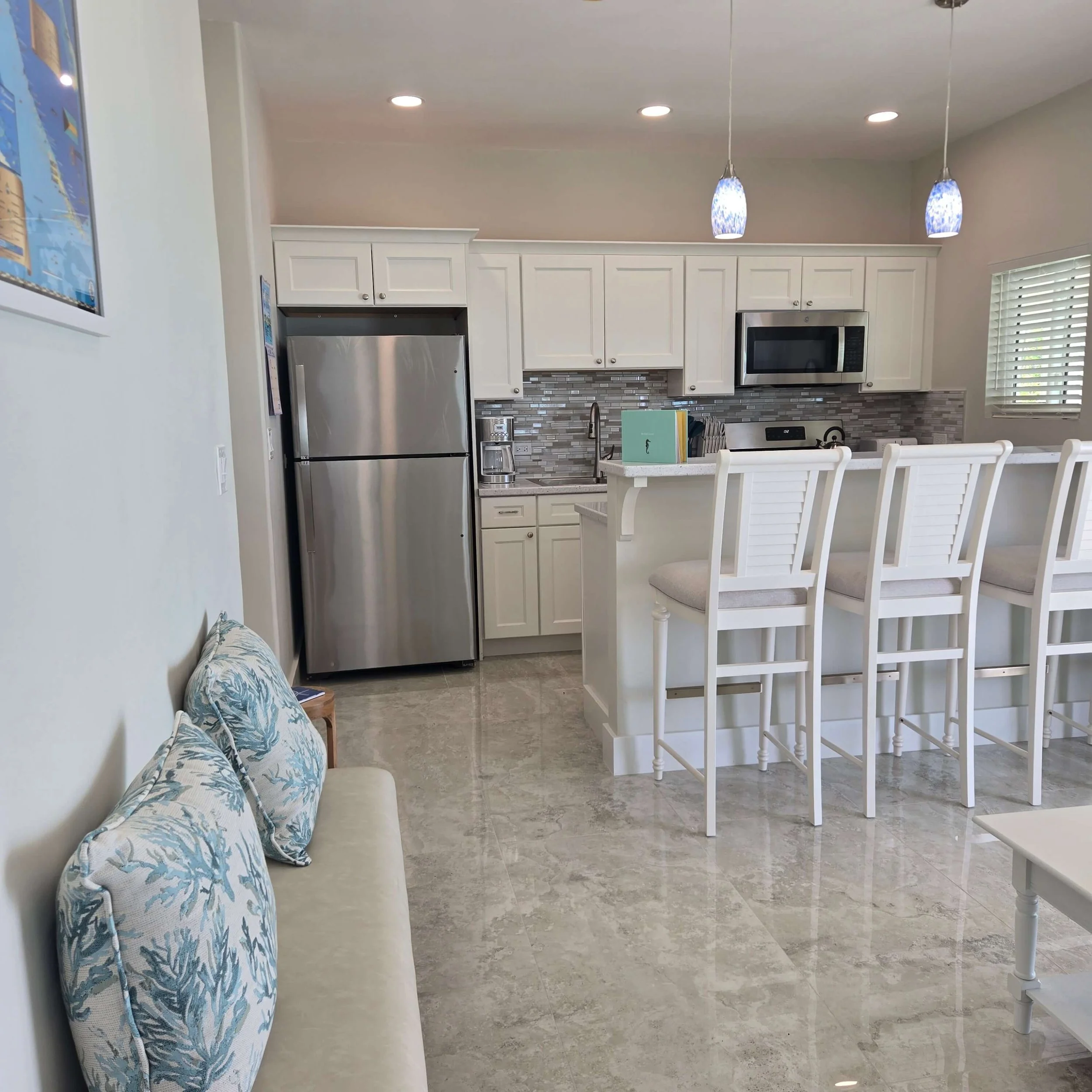 A kitchen with white cabinets, stainless steel appliances, a tiled backsplash, pendant lights hanging from the ceiling, a window with blinds, a counter with a few items including a green book, and a white wooden chair at the counter.