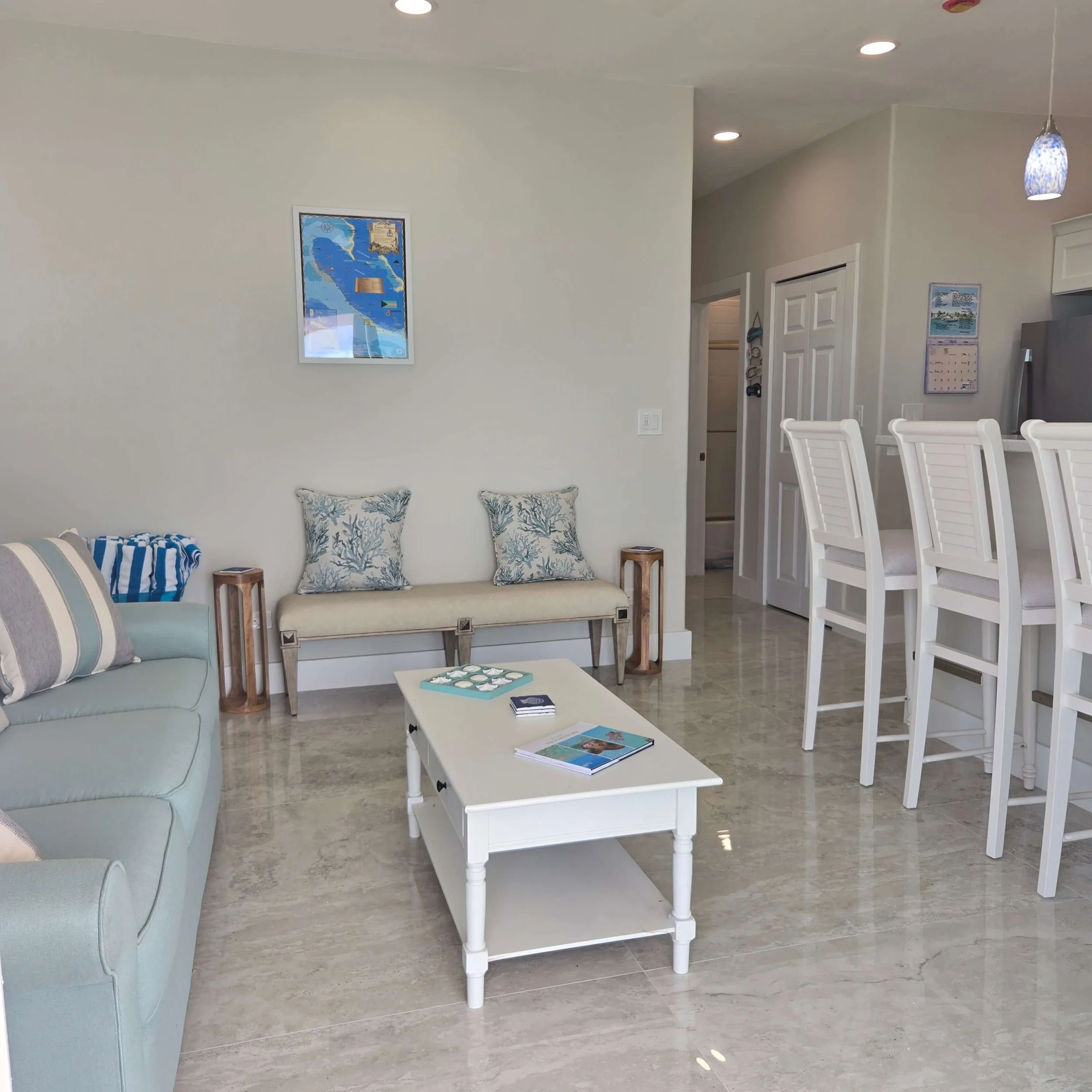 A living room with a white coffee table, beige sofas with blue, white, and coral patterned pillows, white high-back chairs, and wall art including a framed map of Florida.