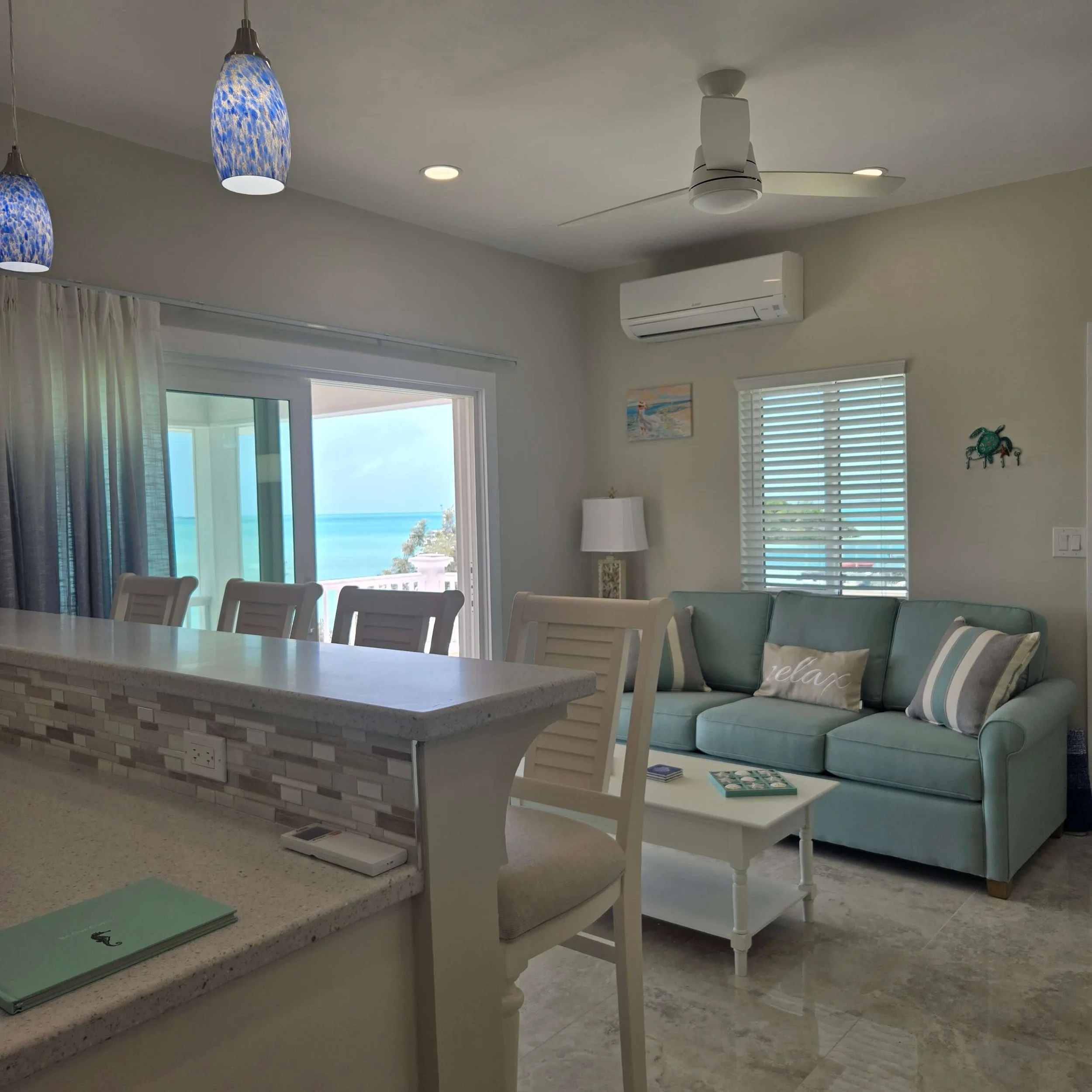 A bright living room with large windows showing ocean views, a white ceiling fan, pendant lights, an air conditioning unit, a light-colored sofa with patterned pillows, a white table, and artwork on the walls.