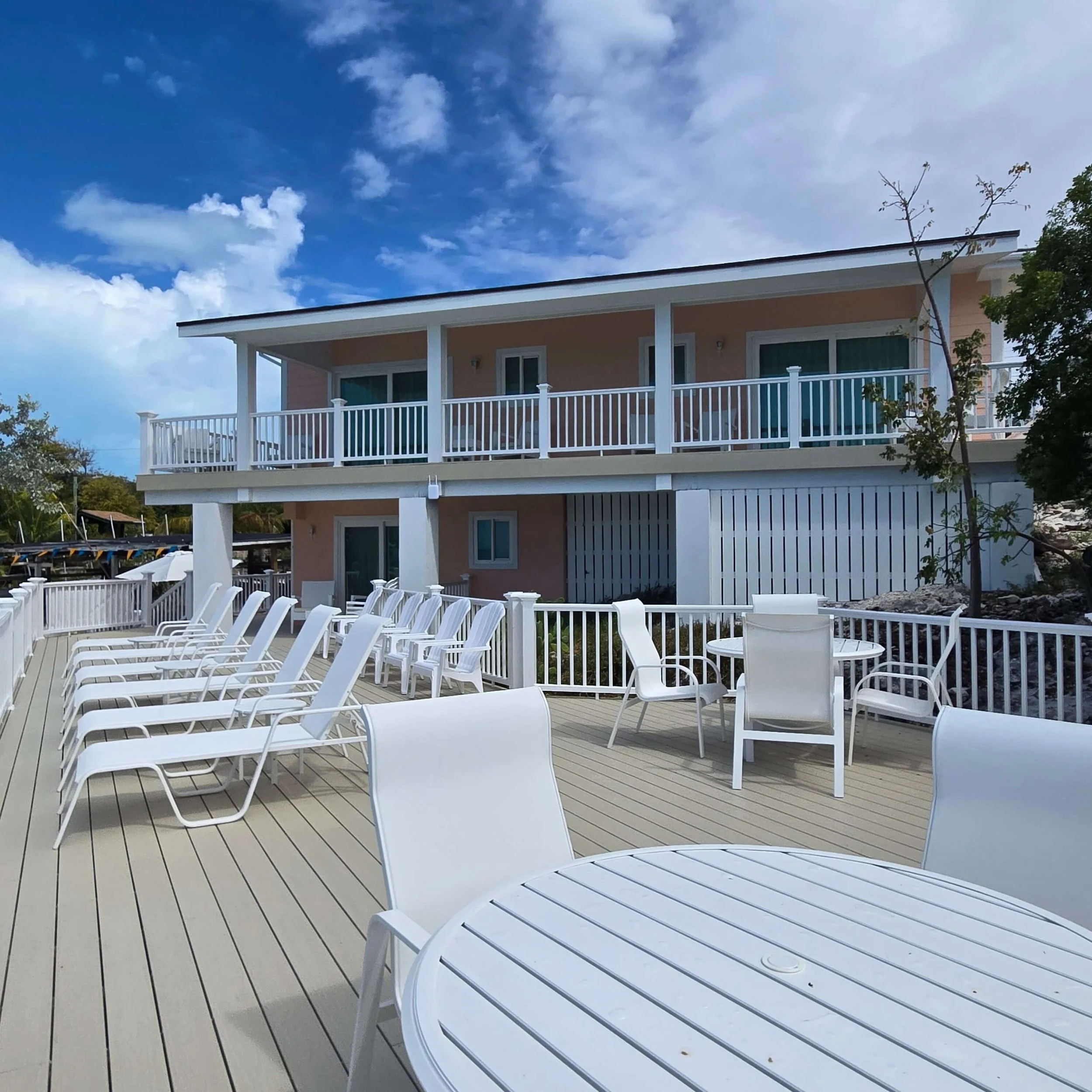 A two-story house with a spacious balcony and a white railing, set against a backdrop of blue sky with scattered clouds. There are white patio chairs on a wooden deck in the foreground, and trees are visible on the side.