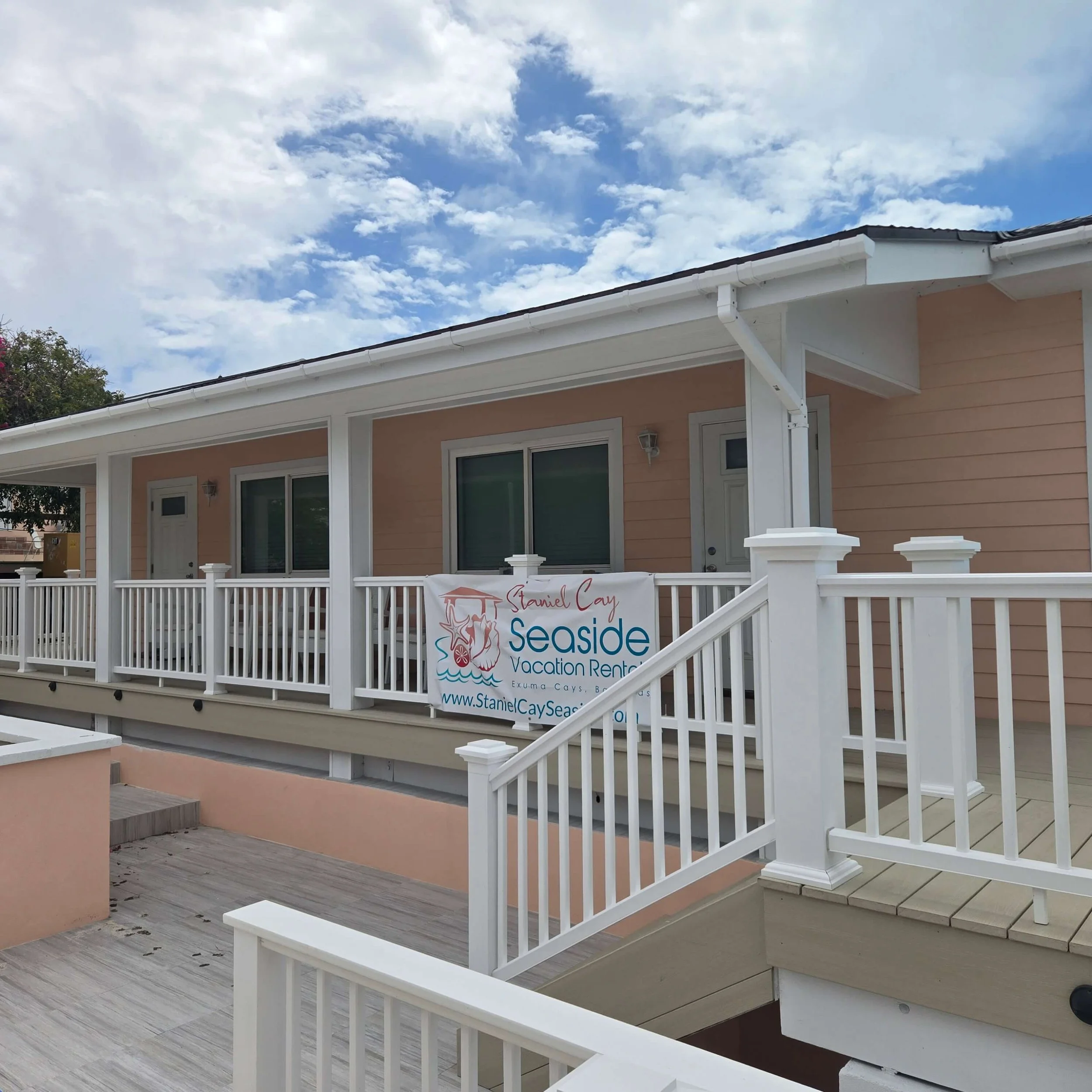 Front porch of a house with white railings and stairs, beige walls, large glass doors, and a sign for a seaside vacation rental. The sky above is partly cloudy.
