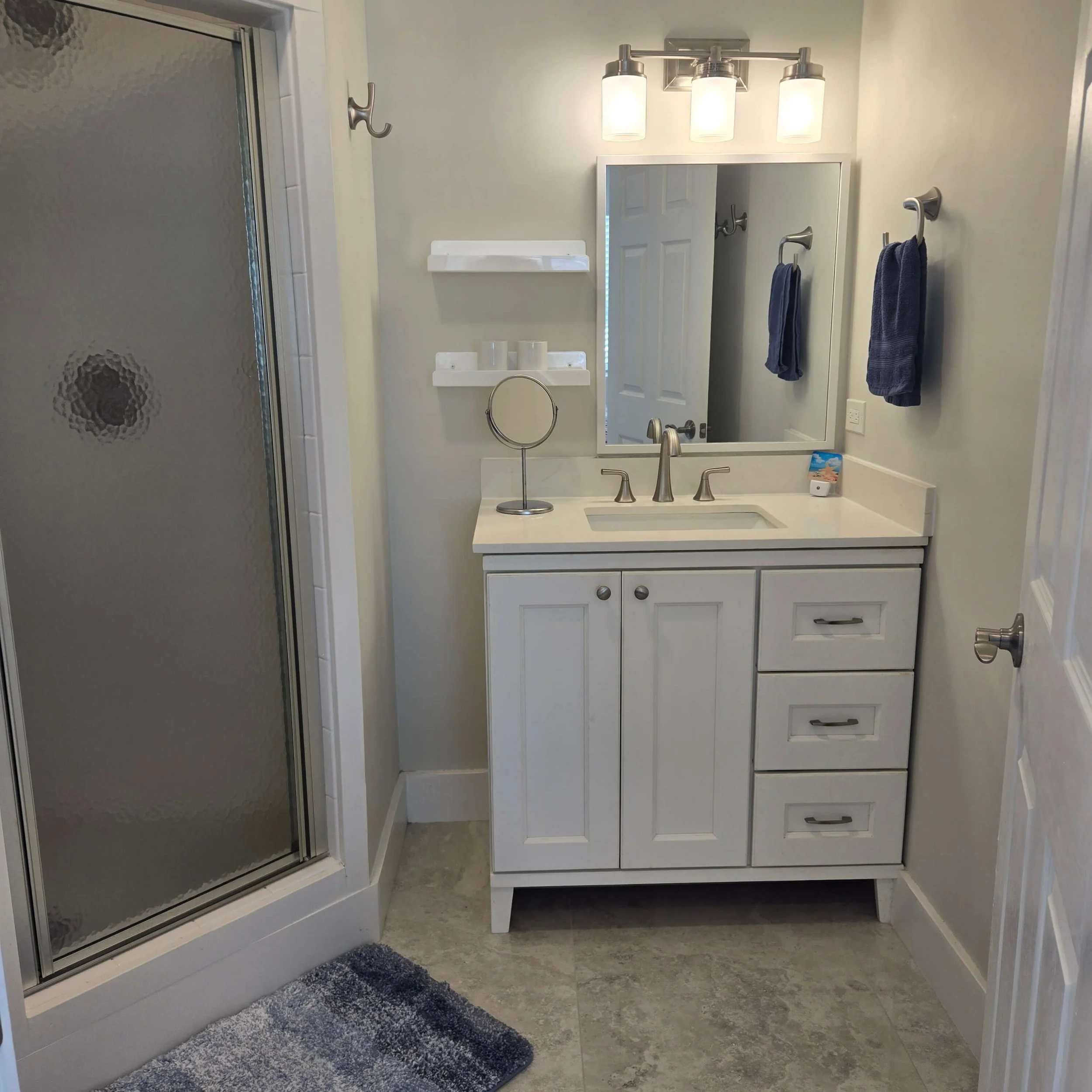 A bathroom vanity with a white cabinet, a large mirror, and three light fixtures above it. Two blue towels hanging on hooks on the wall, a small round mirror, and a gray shaggy rug on the floor. Part of a shower with textured glass door to the left.