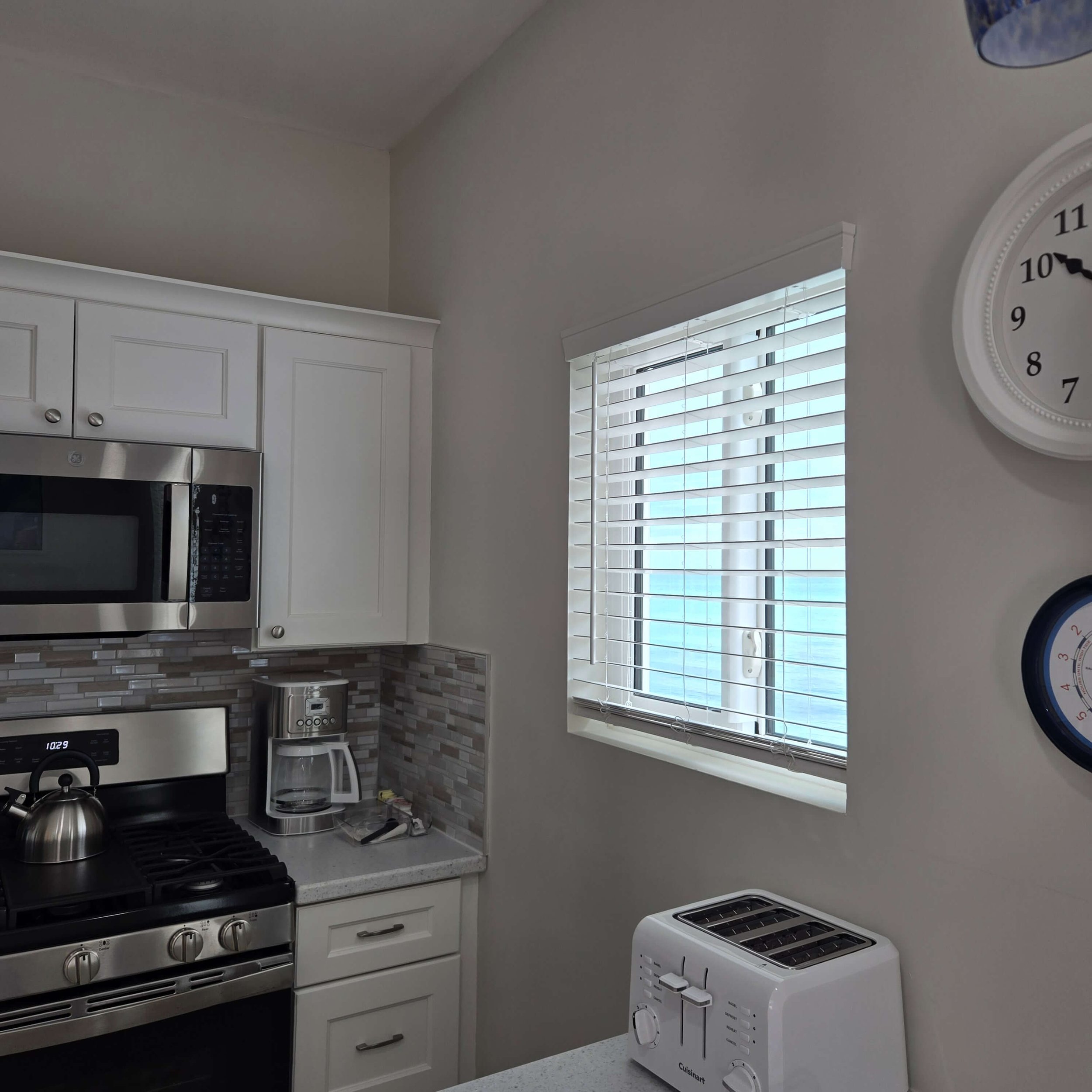A kitchen with white cabinets, stainless steel oven, microwave, and toaster. There is a window with white blinds and a round clock on the wall.