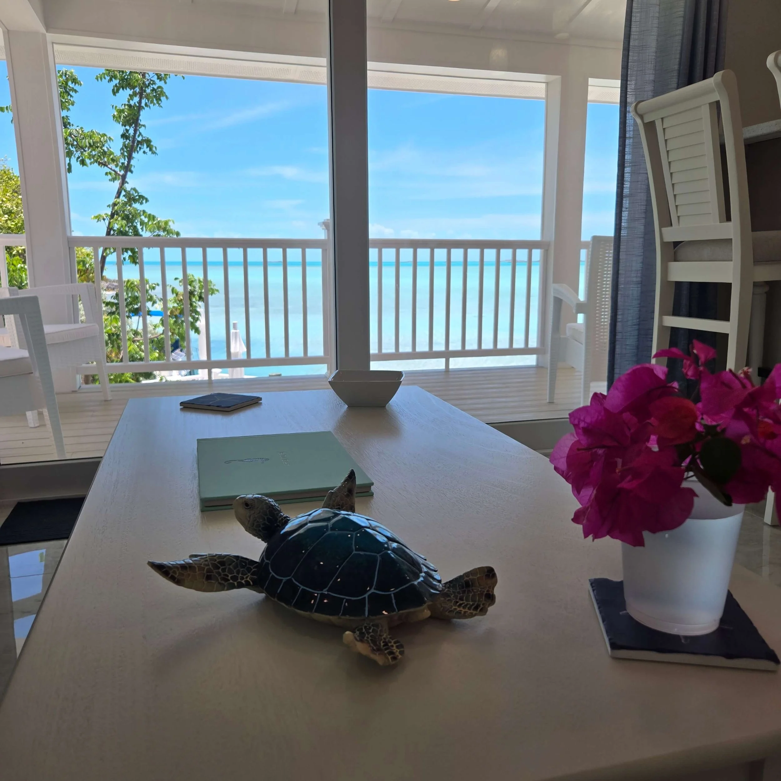 View through glass sliding doors showing a balcony with white railings, green trees, and a clear blue sky with some clouds. Inside, there's a white table with a green notebook, a white bowl, a decorative turtle, and pink flowers in a white pot.