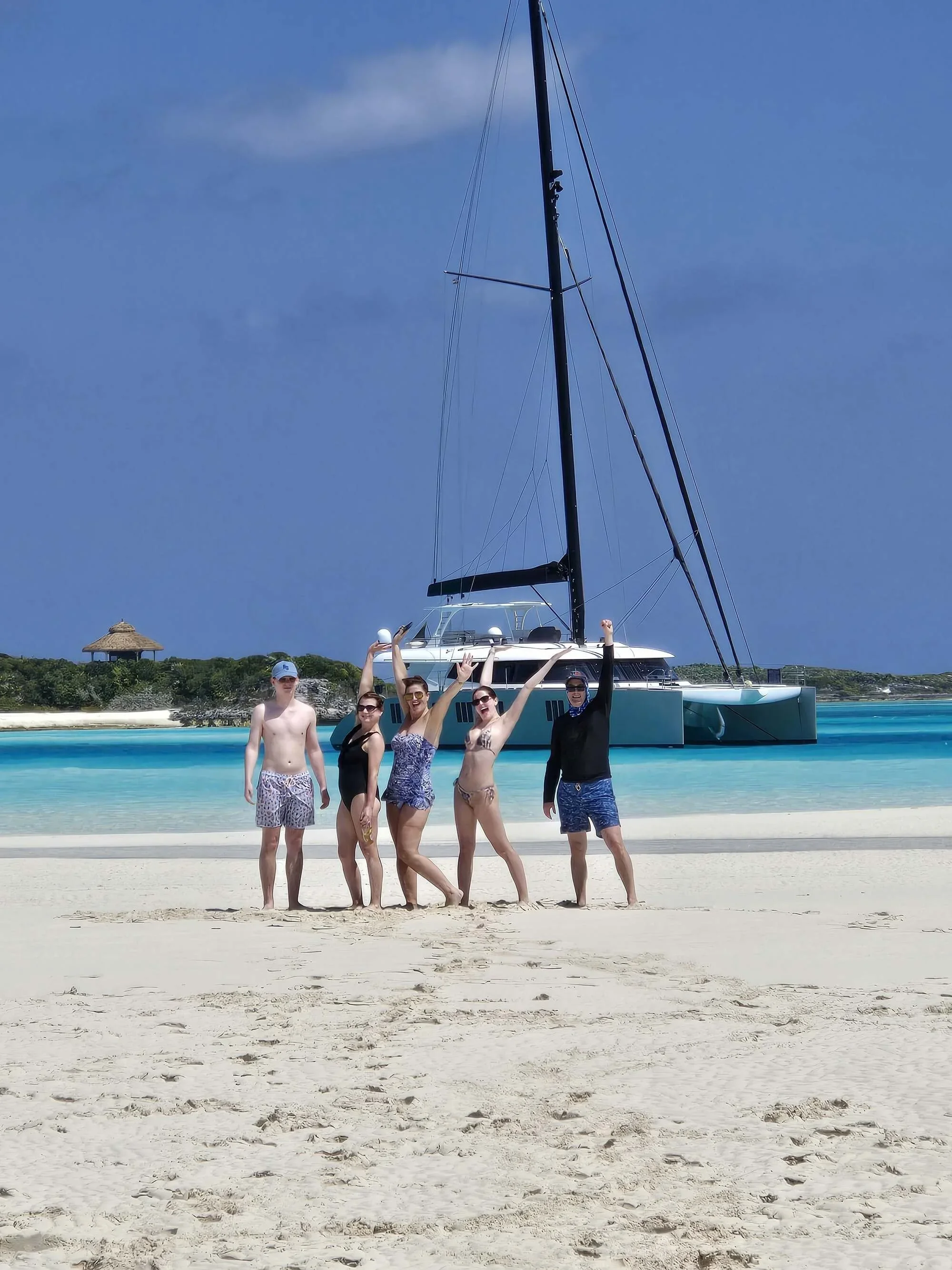 People in swimwear standing on sandy beach in front of a large yacht, with shoreline and a small thatched-roof hut in the background, and blue sky overhead in the Bahamas