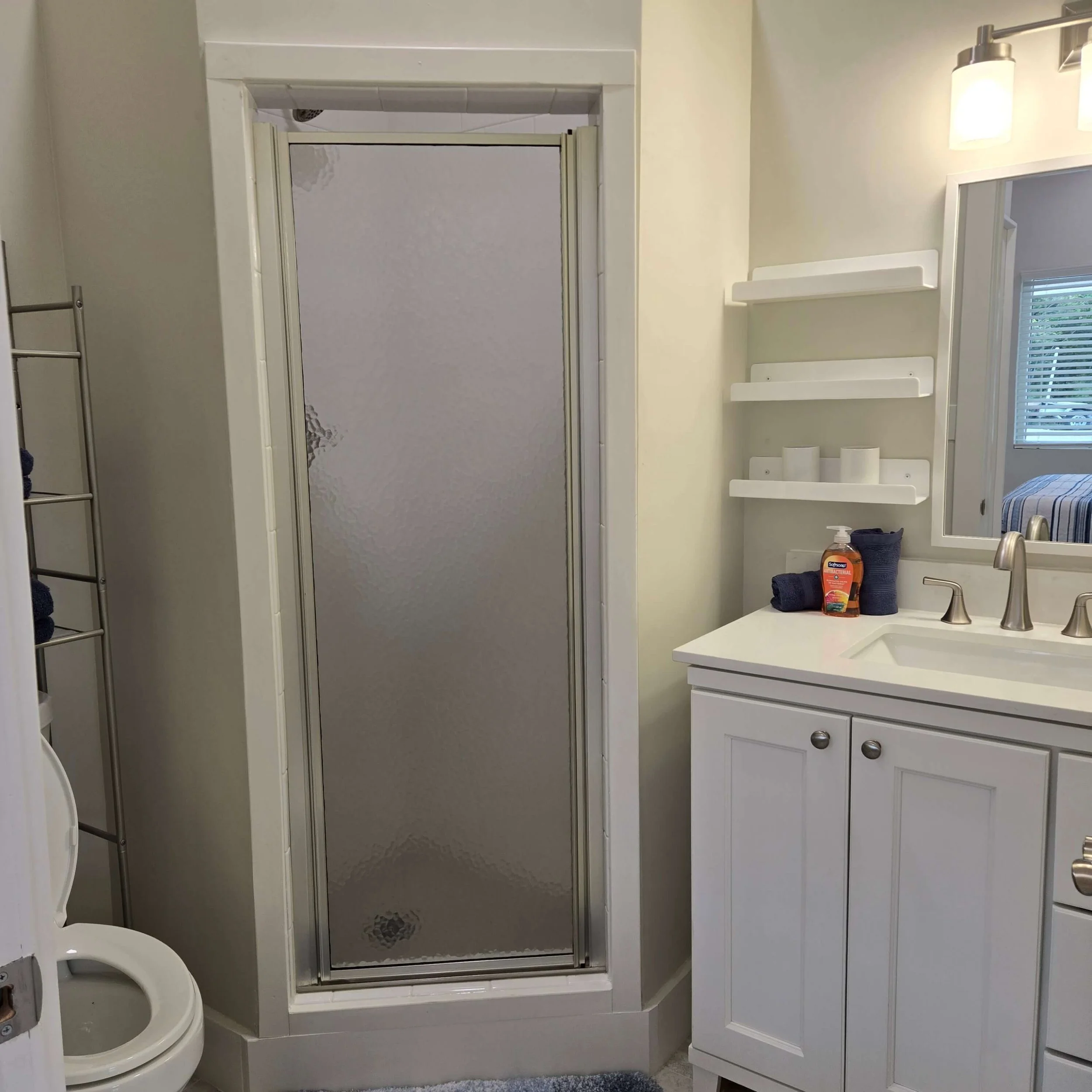A bathroom with a white vanity, a mirror, and a light fixture above the mirror. There is a shower with a sliding door and a towel rack beside it. Towels and toiletries are on the sink.