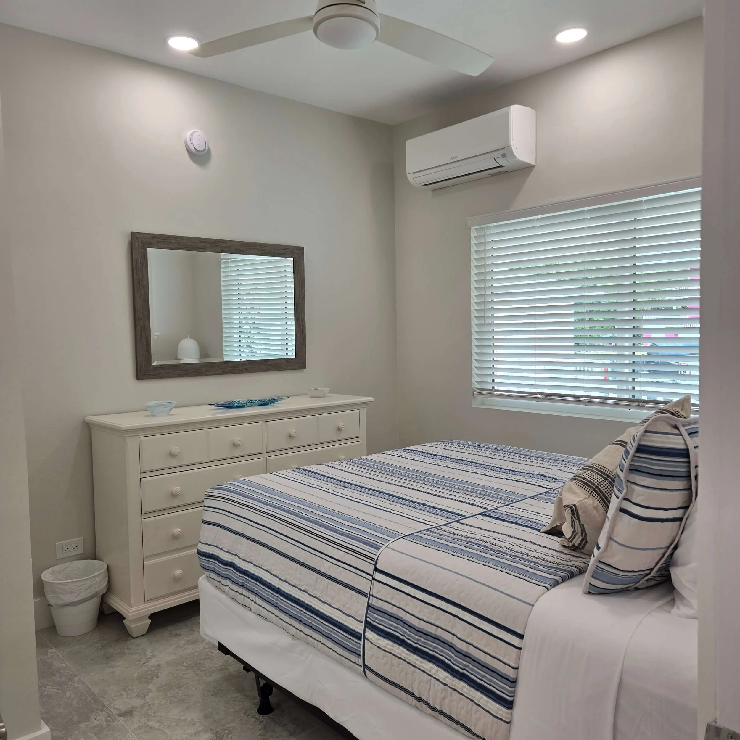 A bedroom with a bed covered in a striped blue, white, and tan comforter, a white dresser with three drawers, a mirror hanging on the wall above the dresser, small bowls on the dresser, a white trash bin on the floor, a window with closed white blind
