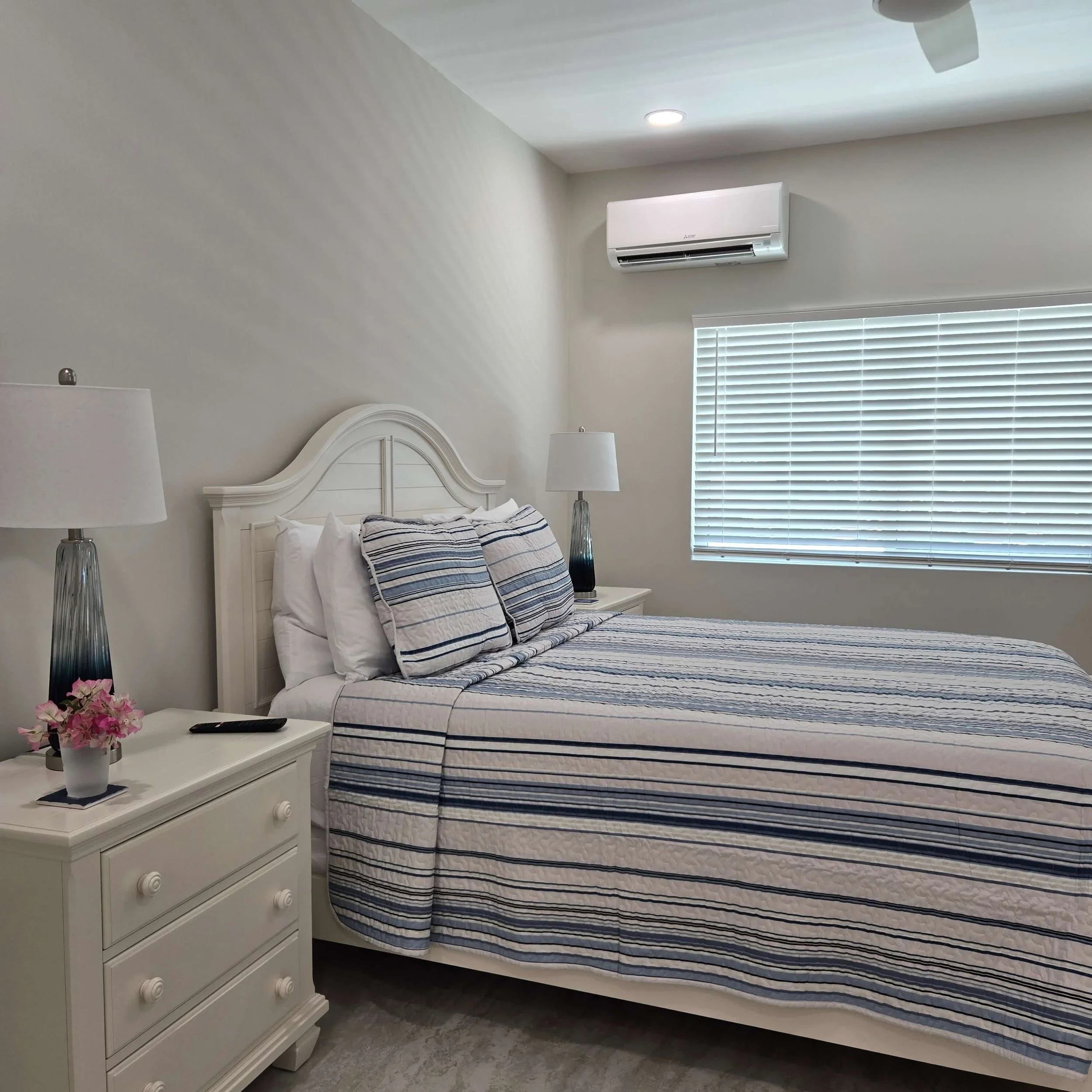 A bedroom with a white wooden bed frame, striped bedding, a white nightstand with a black remote and flowers, two table lamps, window with white blinds, ceiling fan, and wall-mounted air conditioner.