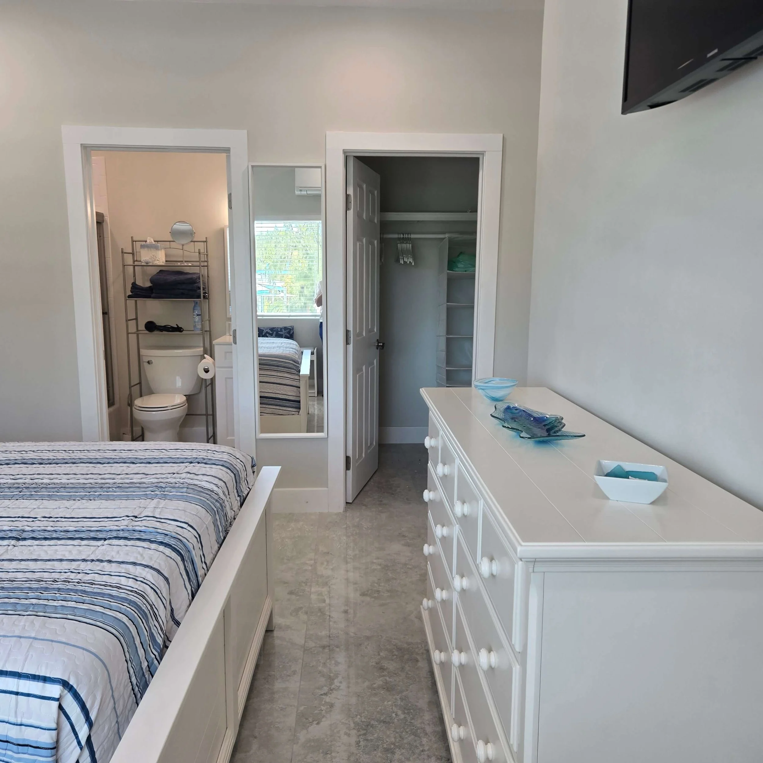 View of a bedroom with a white bed with blue and white striped bedding, a white dresser on the right with blue glass bowls, and a closet with open doors showing shelves. The room has beige tiled flooring, and there are two doorways leading to a small