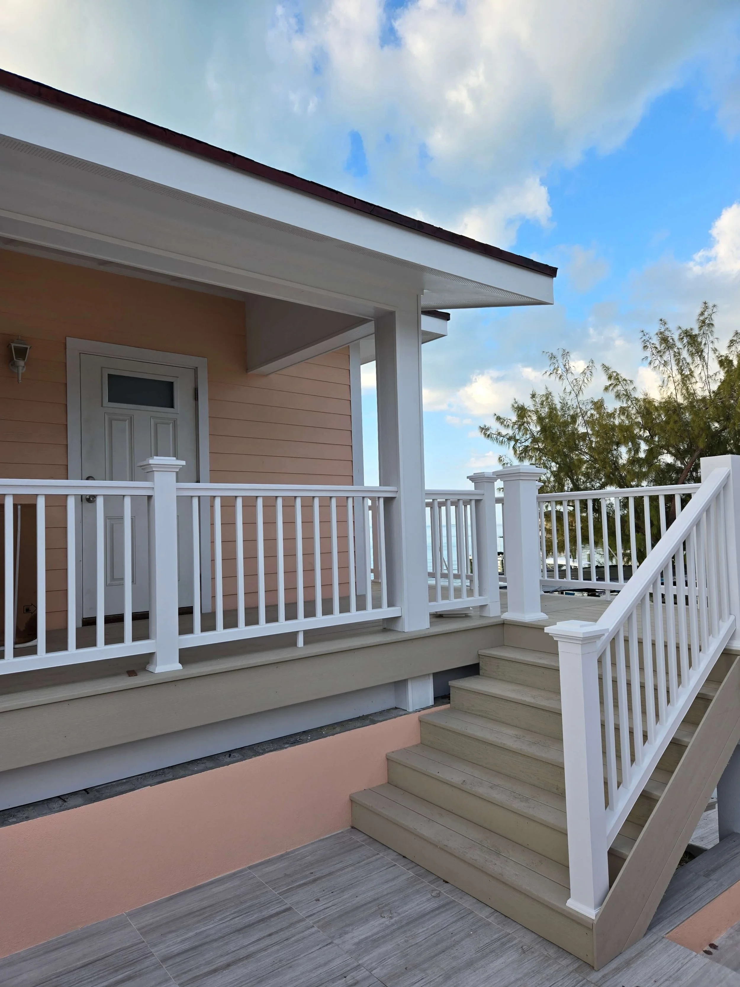 A wooden front porch with white railing, a set of stairs, a peach-colored house wall, a door, and a small wall-mounted light fixture. There are trees and a cloudy sky in the background.