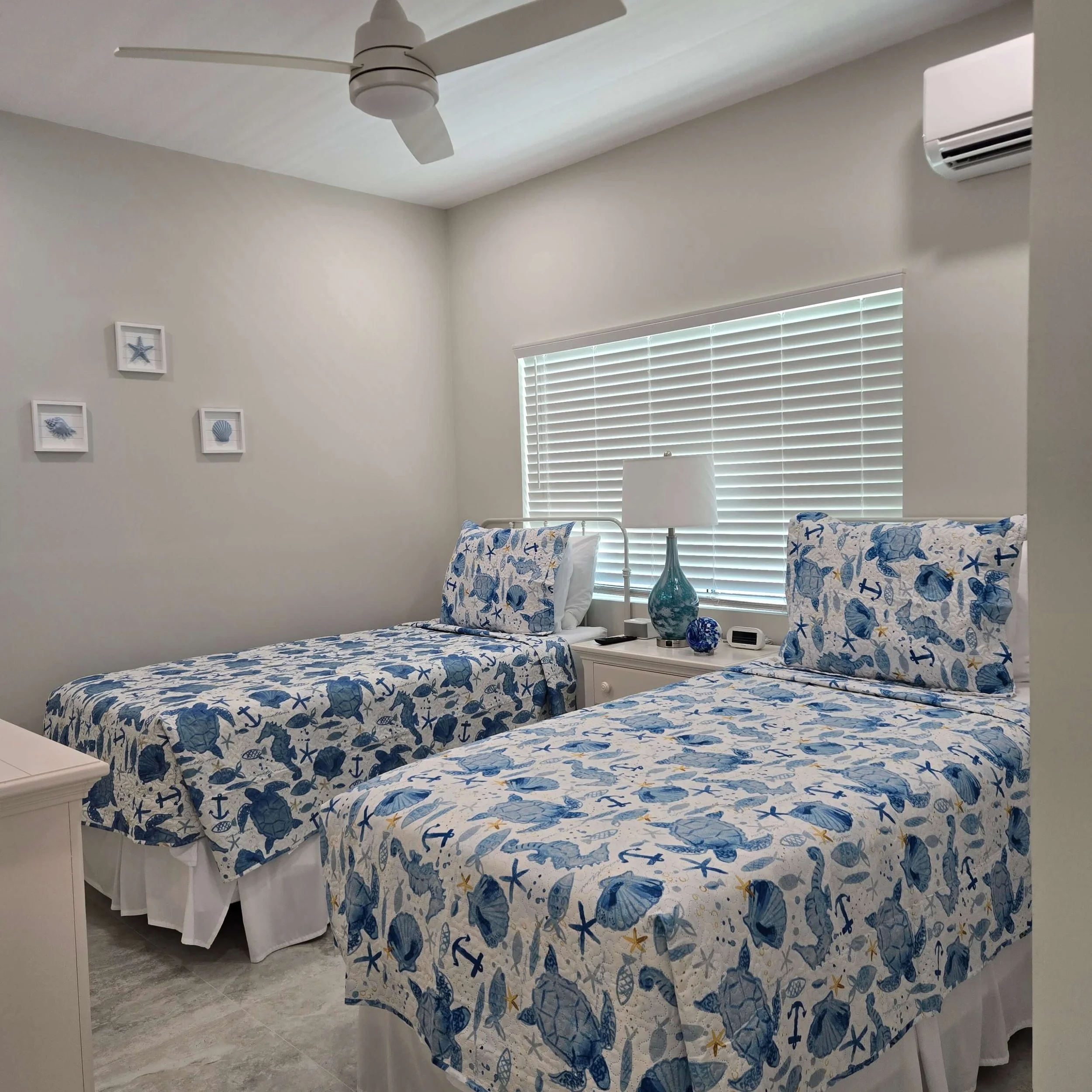 A bedroom with coastal decor featuring two beds with sea-themed bedspreads and pillows, a nightstand with a lamp, wall art with seashells, a window with white blinds, a ceiling fan, and an air conditioning unit.