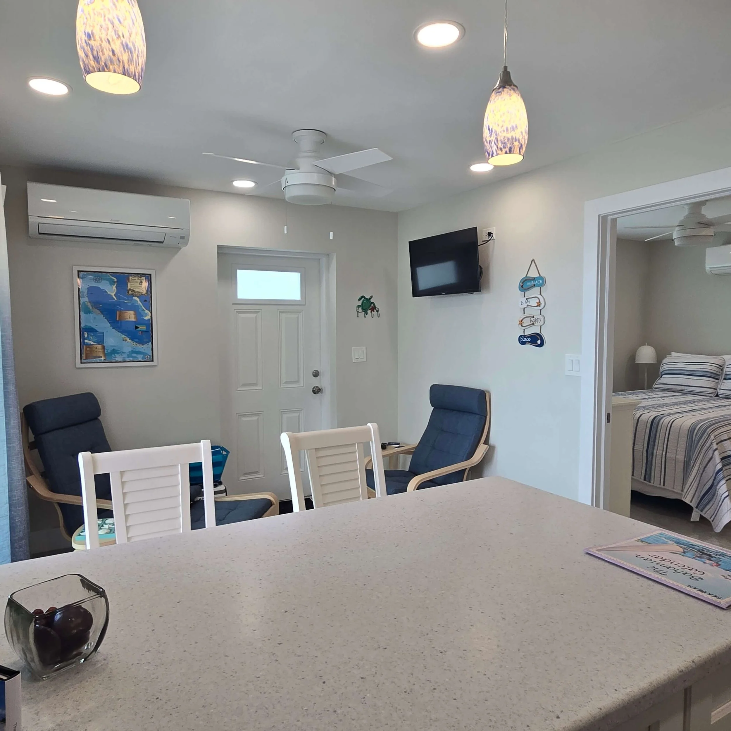 A kitchen or dining area with a white countertop, white chairs, a bowl of cherries, a glass of wine, and documents or magazines on the counter. Background includes a white door, framed picture, TV, and hanging pendant lights.