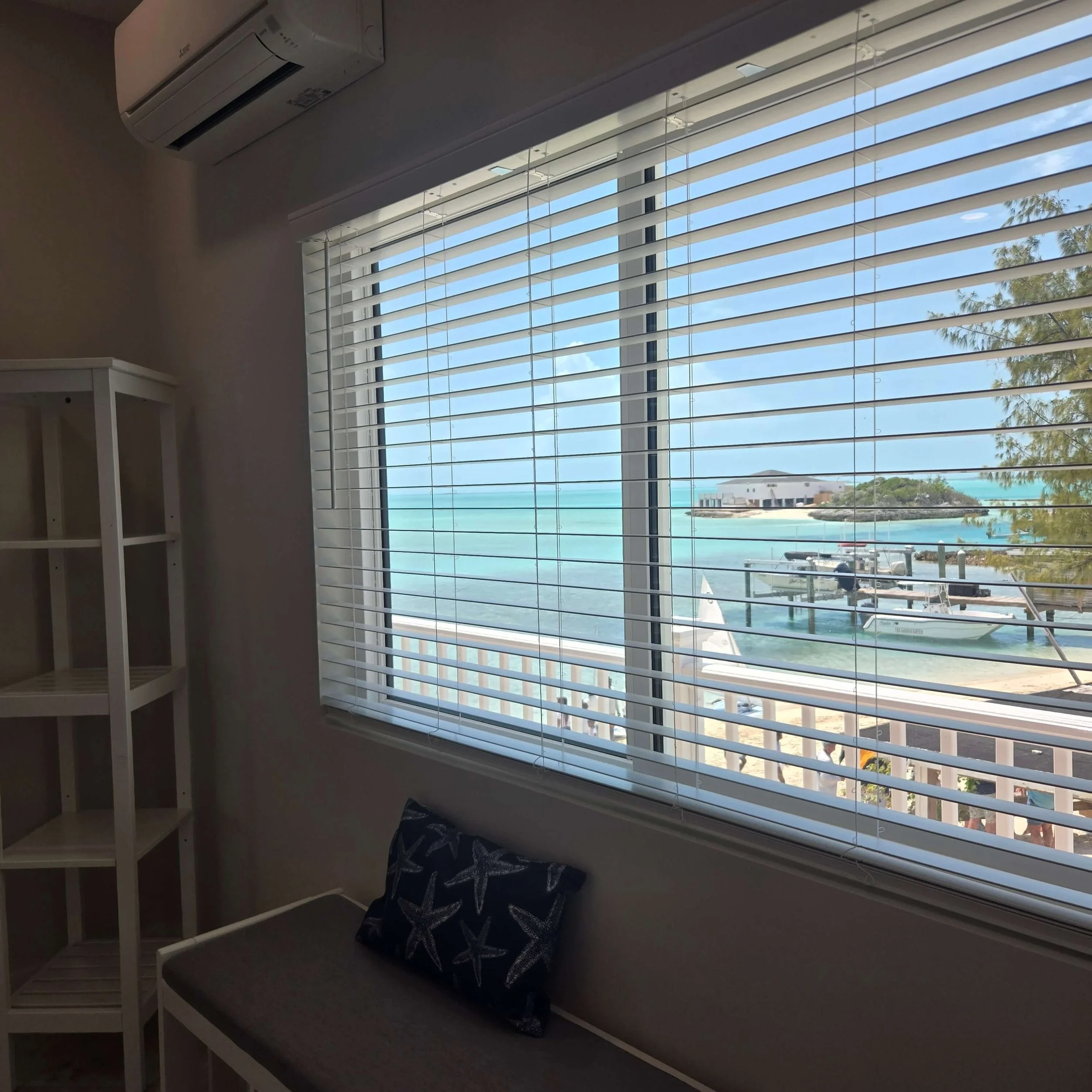 A room with a large window with white blinds, overlooking an ocean view with sandy beach and trees. There is a white bookshelf on the left and a cushioned bench or window seat with a pillow in front of the window.