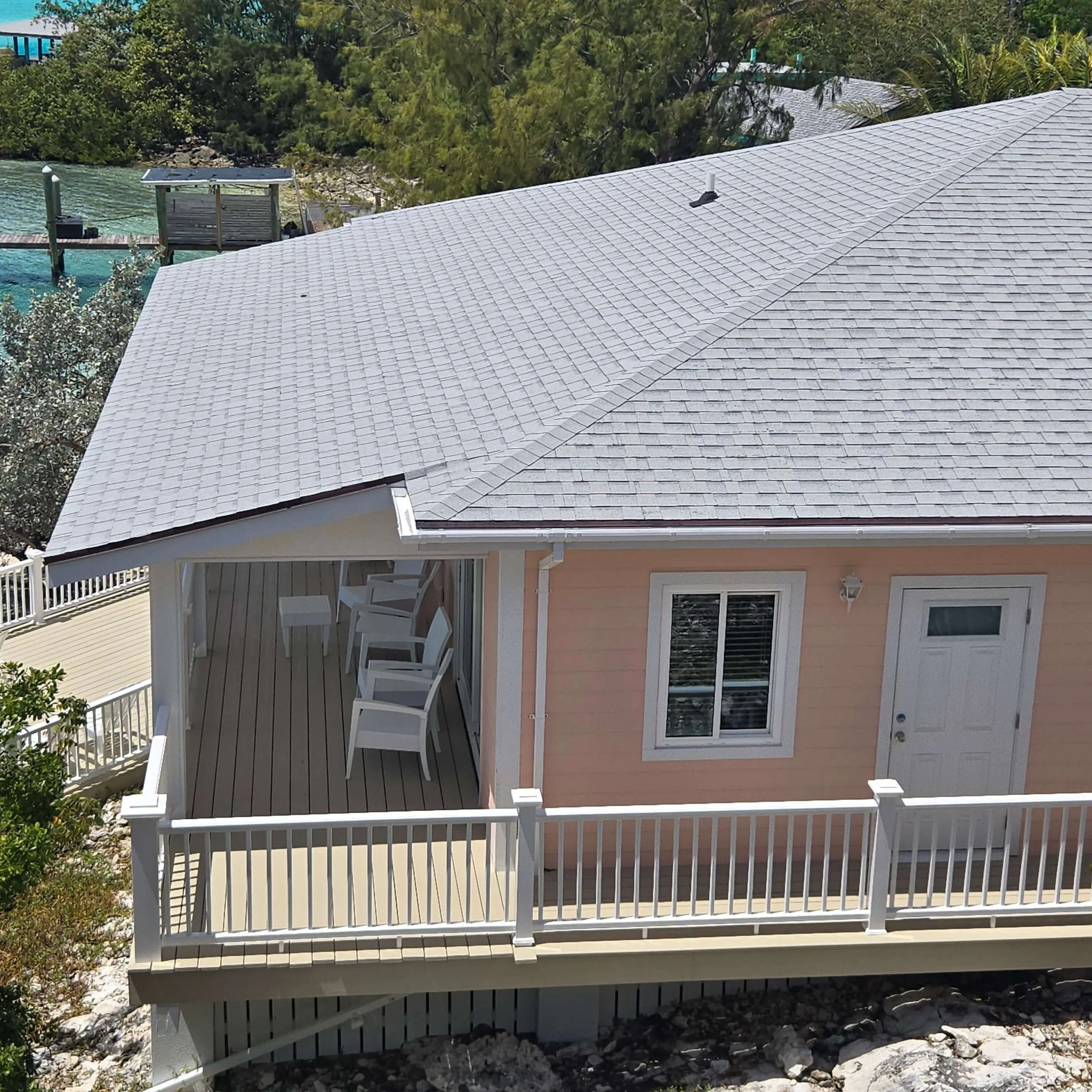 A house with a gray shingle roof, a small white porch with chairs, and a white railing, with trees and water in the background.