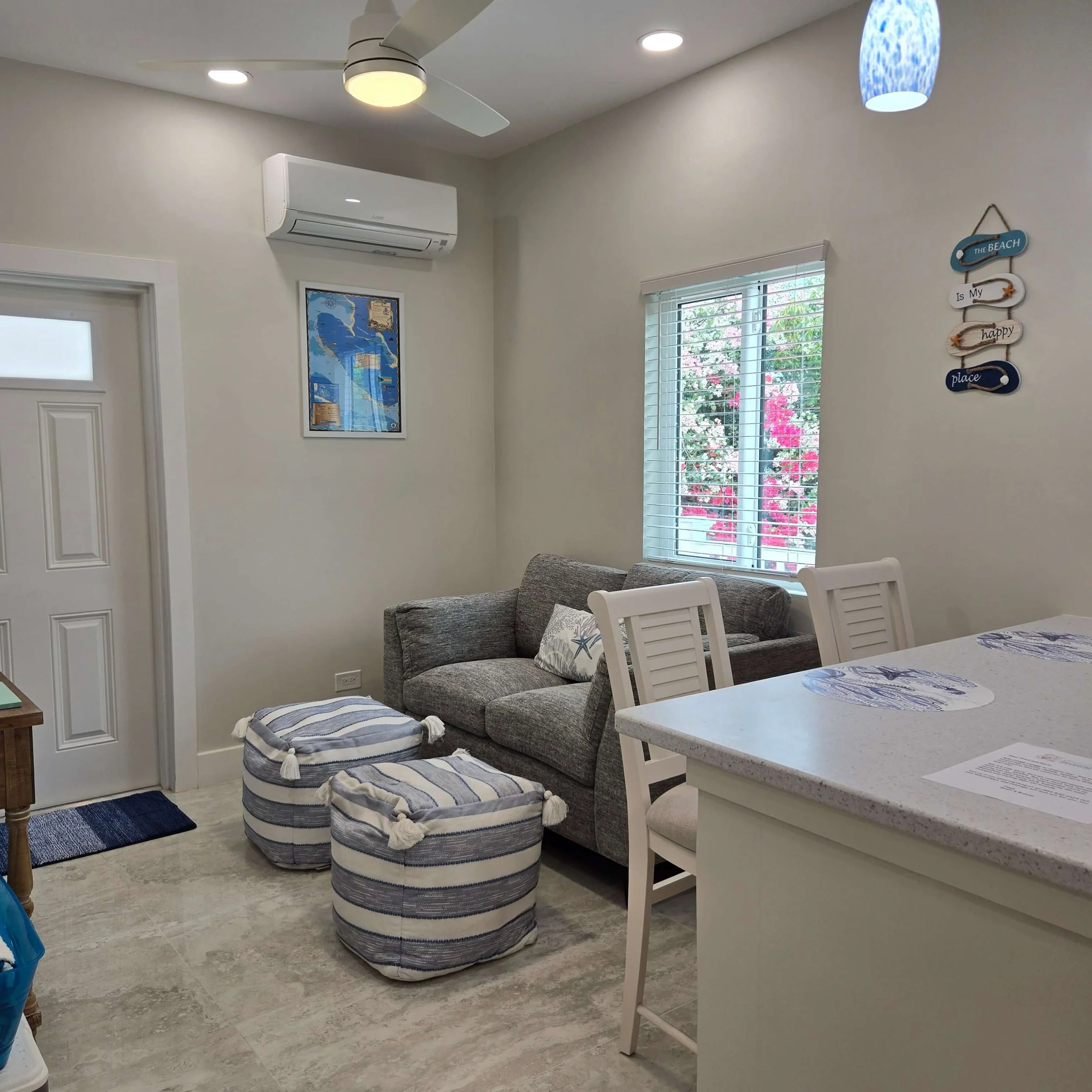 Interior of a living room with a ceiling fan, air conditioner, framed map on the wall, a window with blinds, a blue hanging light, and a wall decoration reading "The beach is my happy place." A gray sofa with patterned pillows and a striped ottoman a