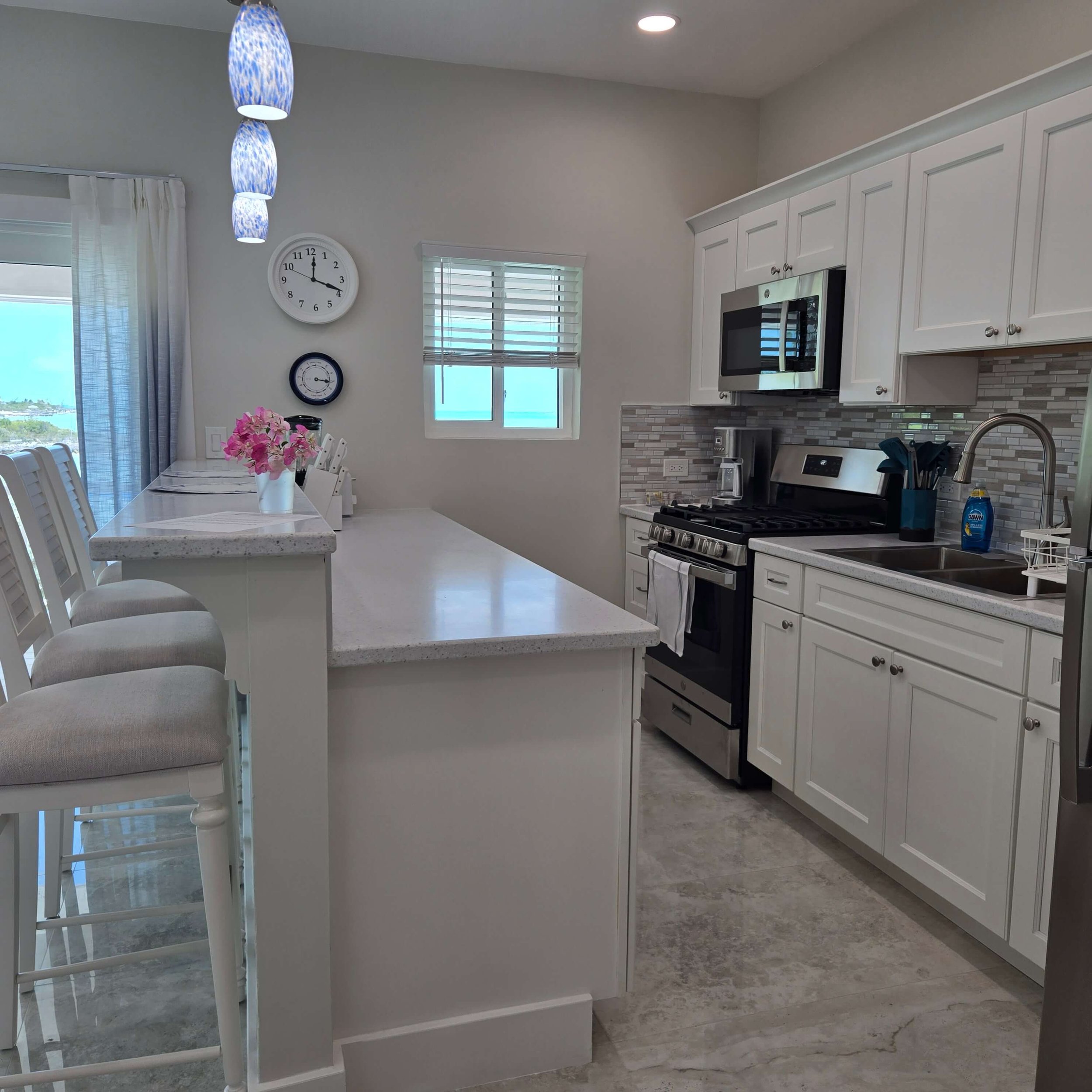 Kitchen with white cabinets, stainless steel microwave, stove, and backsplash, with a white table and chairs, pink flowers, and a view outside through the windows.