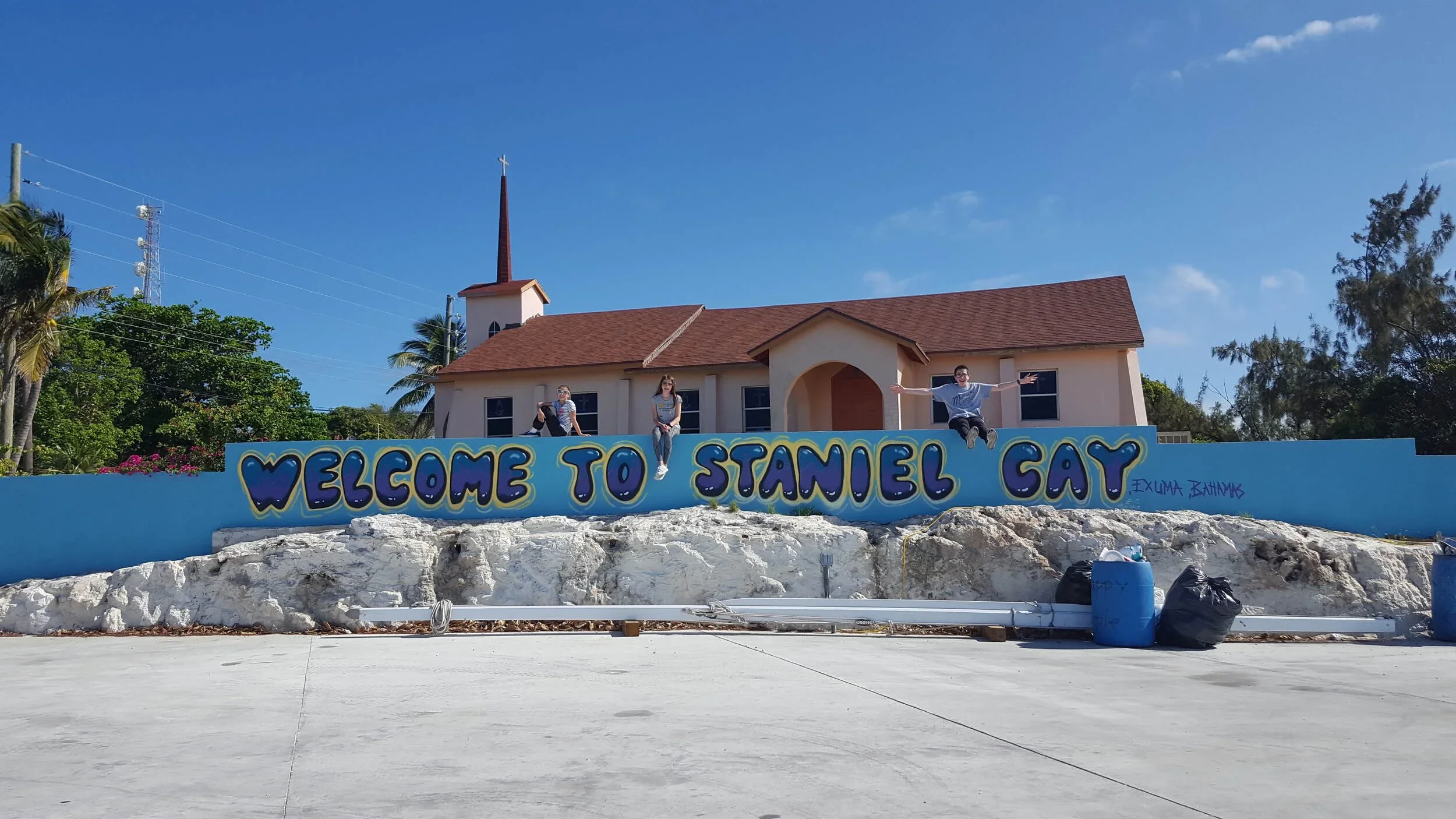A group of three people sitting on a colorful sign that says 'Welcome to Staniel Cay' in front of a church building with a steeple, palm trees, and clear blue sky.