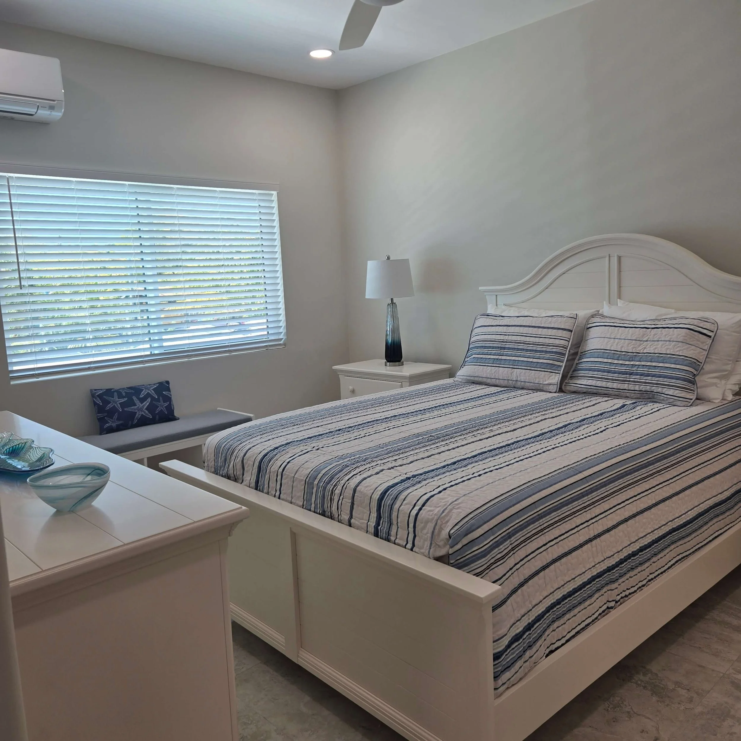 A bedroom with a white bed frame, striped bedding, a bedside table with a lamp, window with blinds, and a ceiling fan.