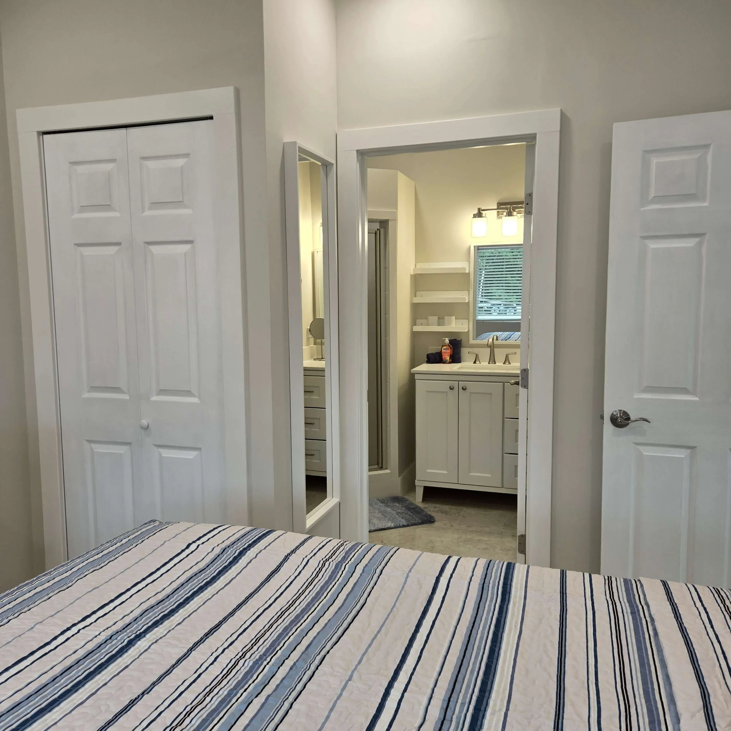 View of a bedroom with a bed in the foreground with a striped bedspread, an open door revealing a bathroom with a vanity and mirror, and a closet with white double doors.