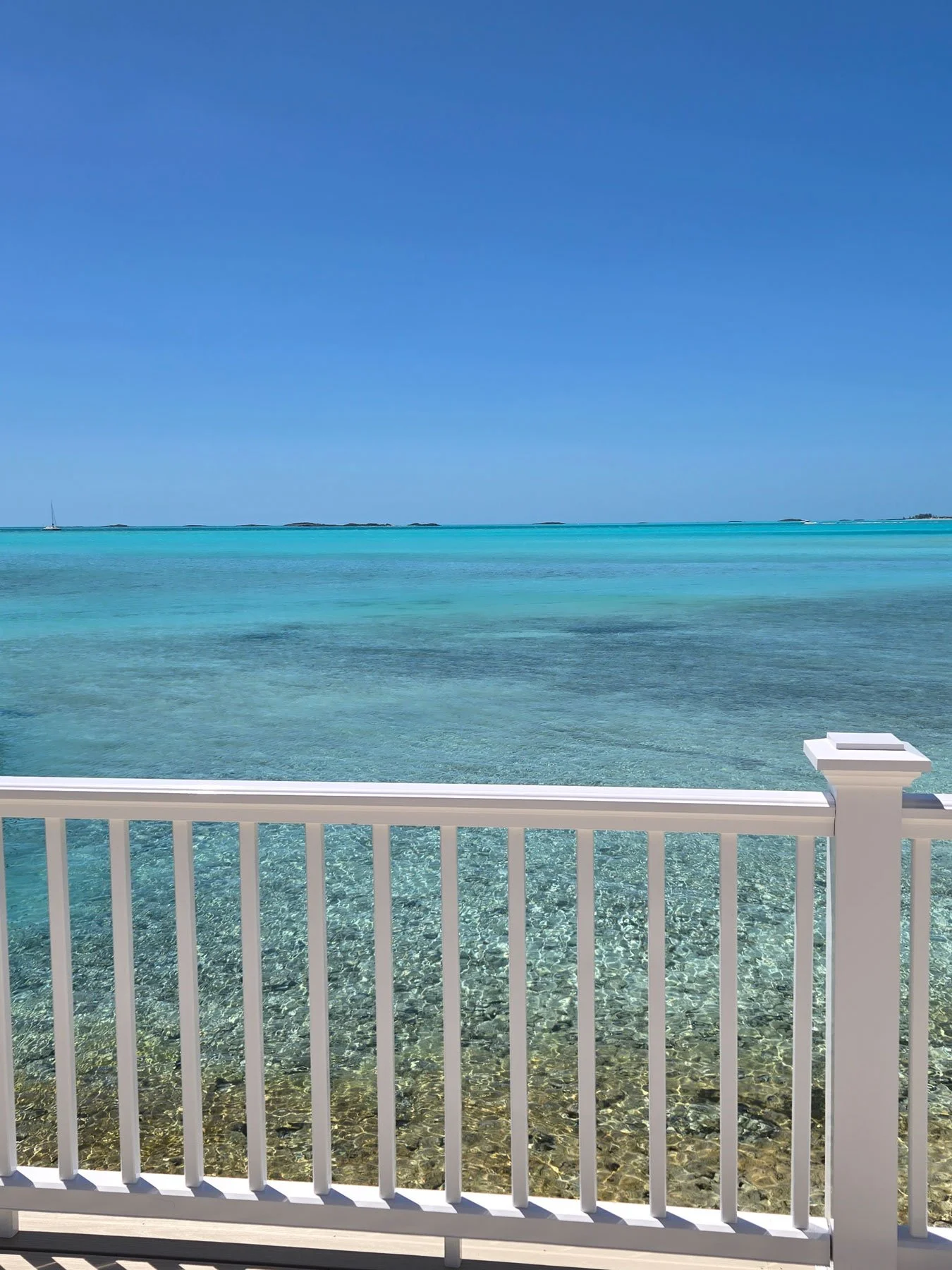 View of clear turquoise ocean water seen from a balcony with a white railing. In the distance, a sailboat is on the horizon, and there are small islands or rocks. The sky is bright blue with no clouds.