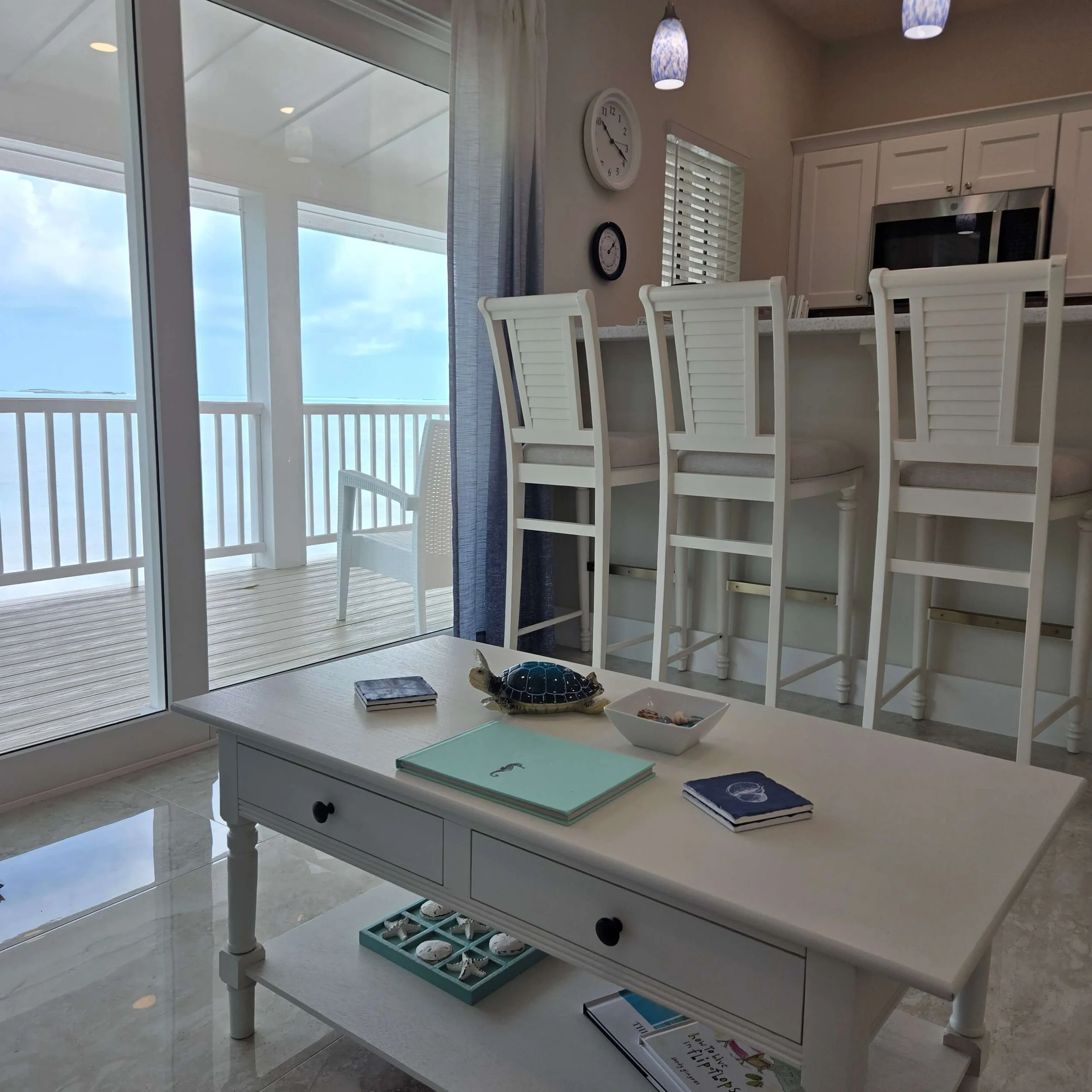A white wooden dining table with built-in shelves, holding books, shells, and decorative items, situated in a bright room with large windows and sliding glass doors leading to an outdoor balcony with a view of the ocean or sky.