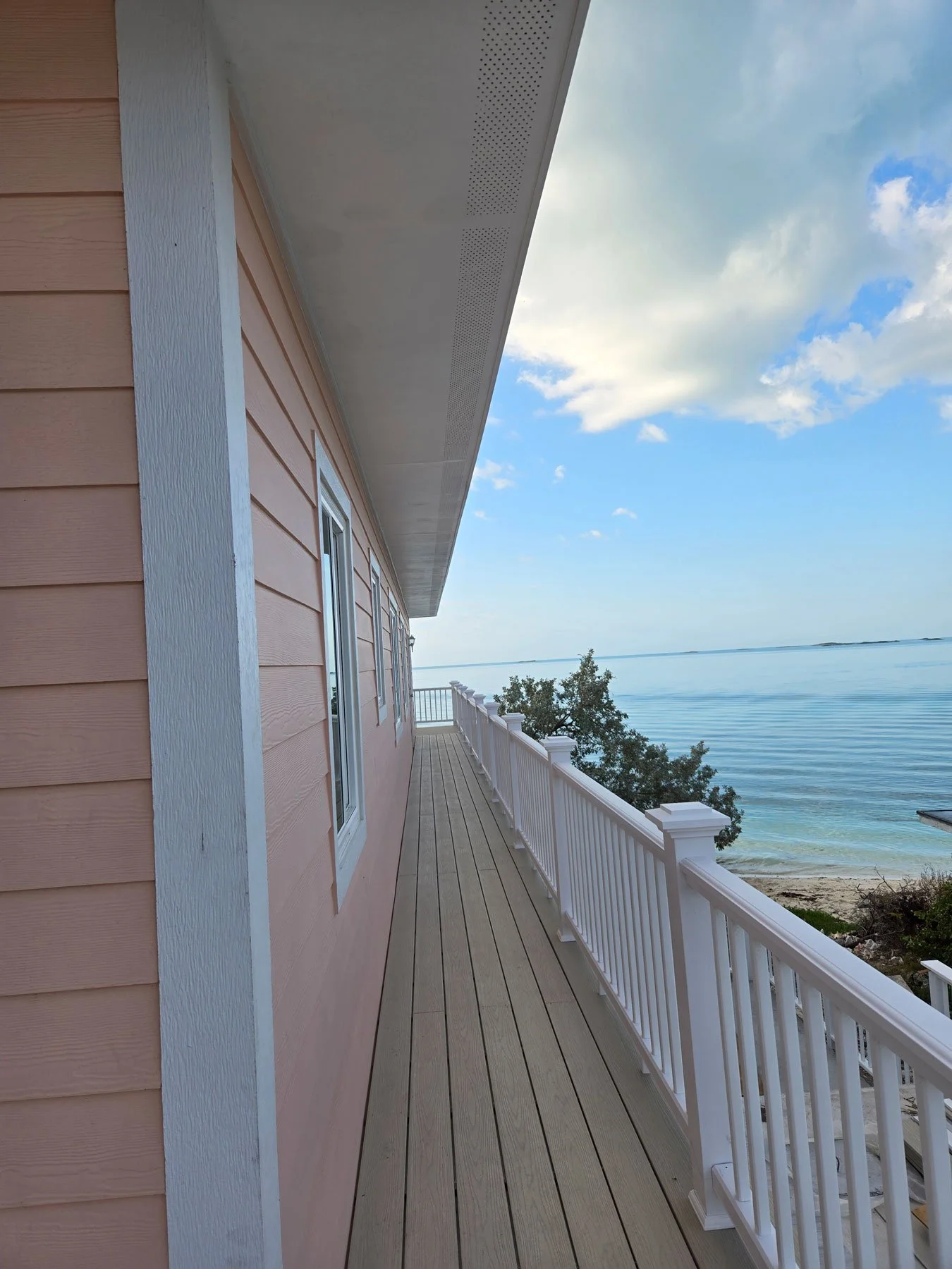 View of a beachfront house with pink siding, white trim, and a wooden deck, overlooking calm water and partly cloudy skies.