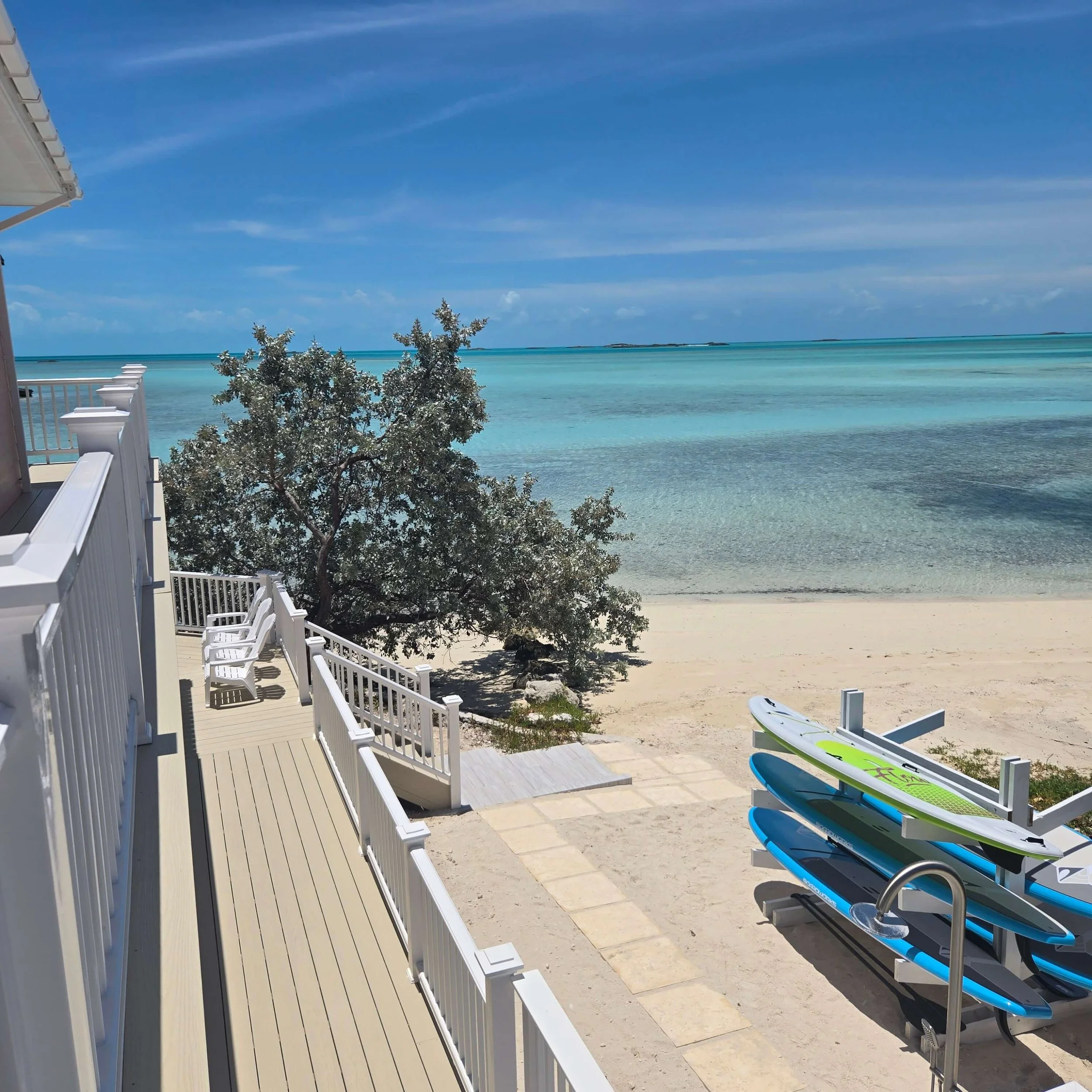 Seaside's beachside deck with white railings and furniture, overlooking clear turquoise water and a distant island under a blue sky with a few clouds. SUPs are stacked on the beach for guest use.